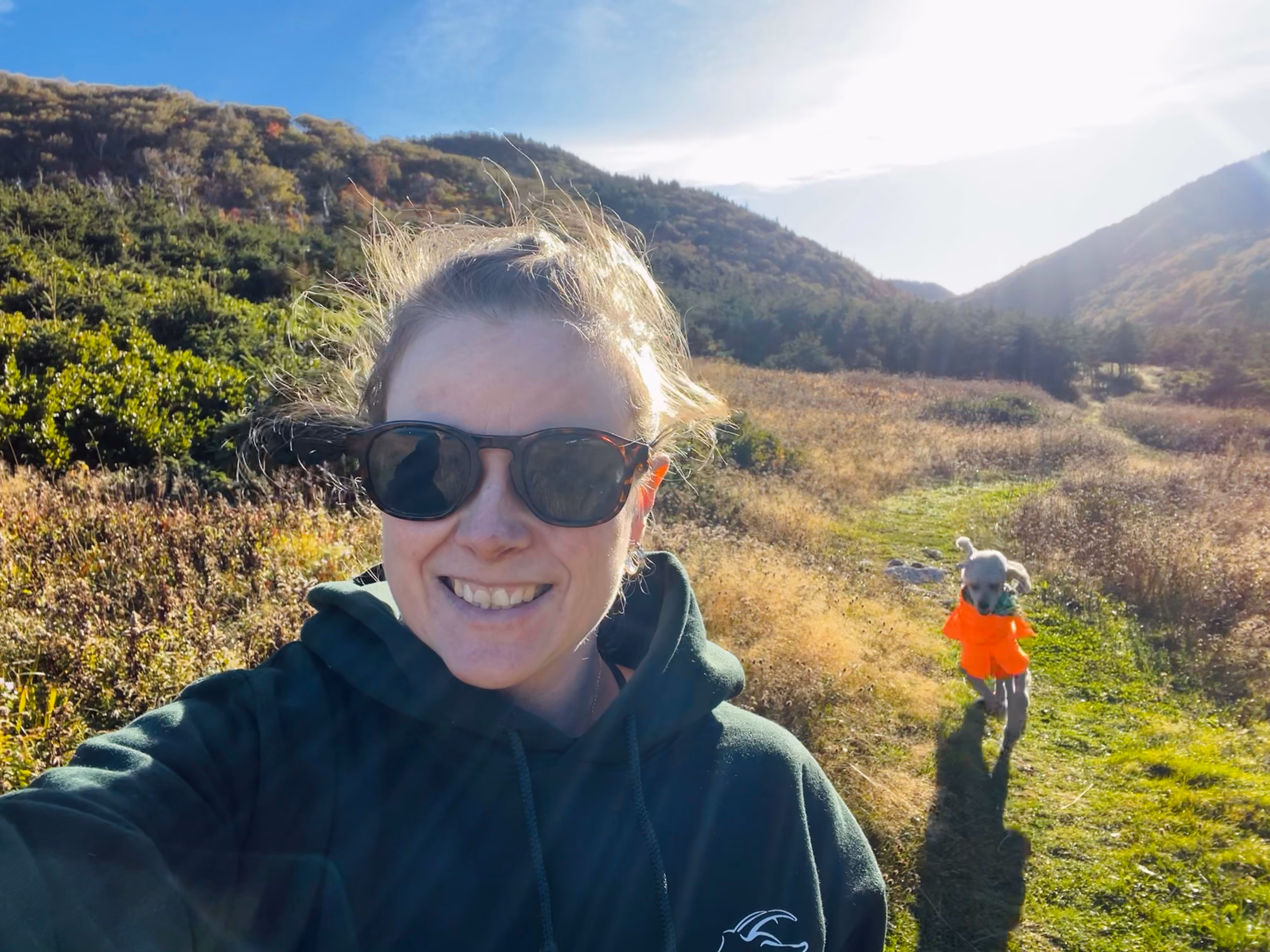 Jess smiles in a selfie on a sunny hiking trail through autumn hills, her poodle in an orange coat running along the path behind her.