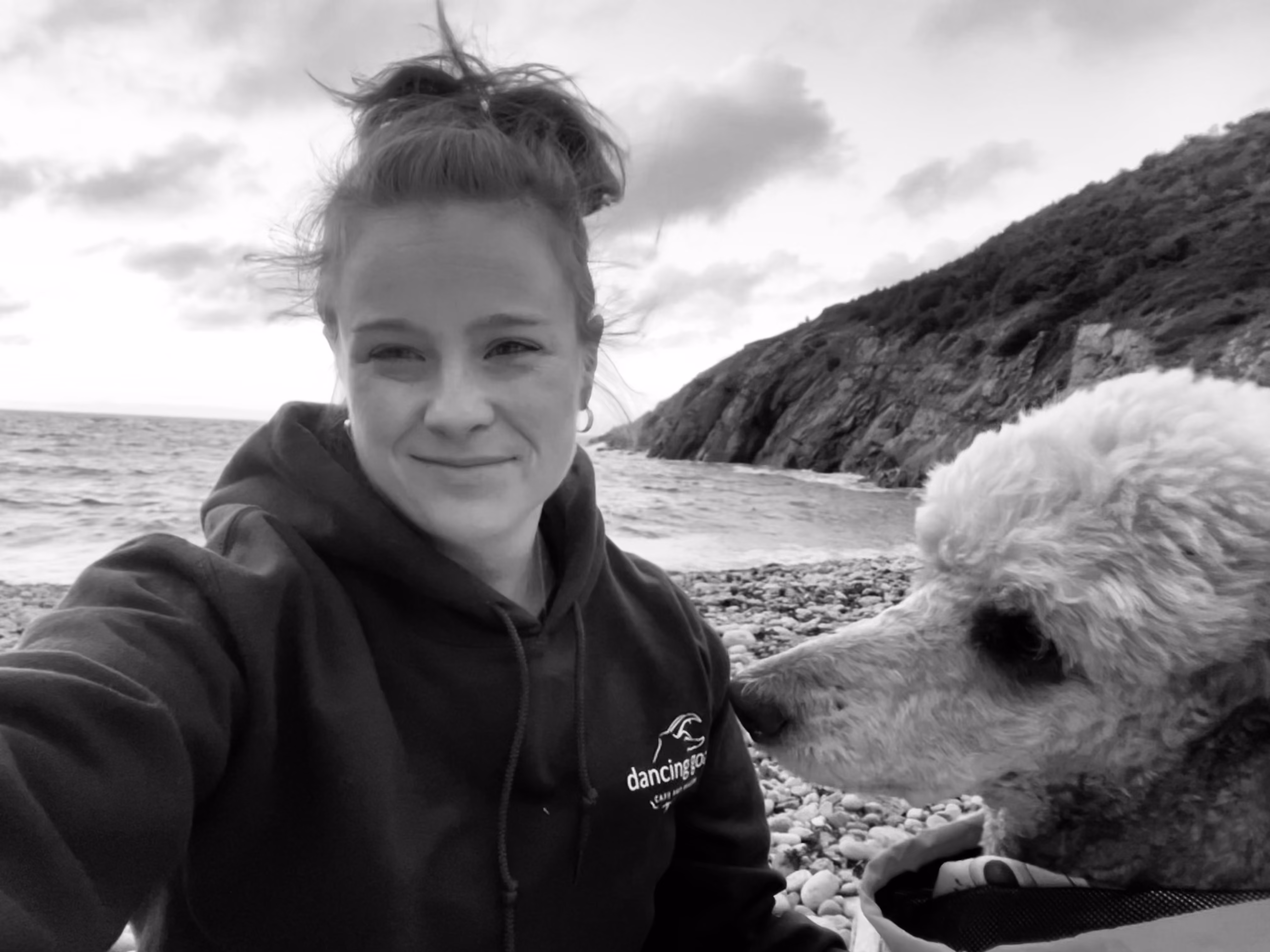Black-and-white selfie of Jess on a pebble beach with her poodle beside her, the ocean and a rocky headland behind them.