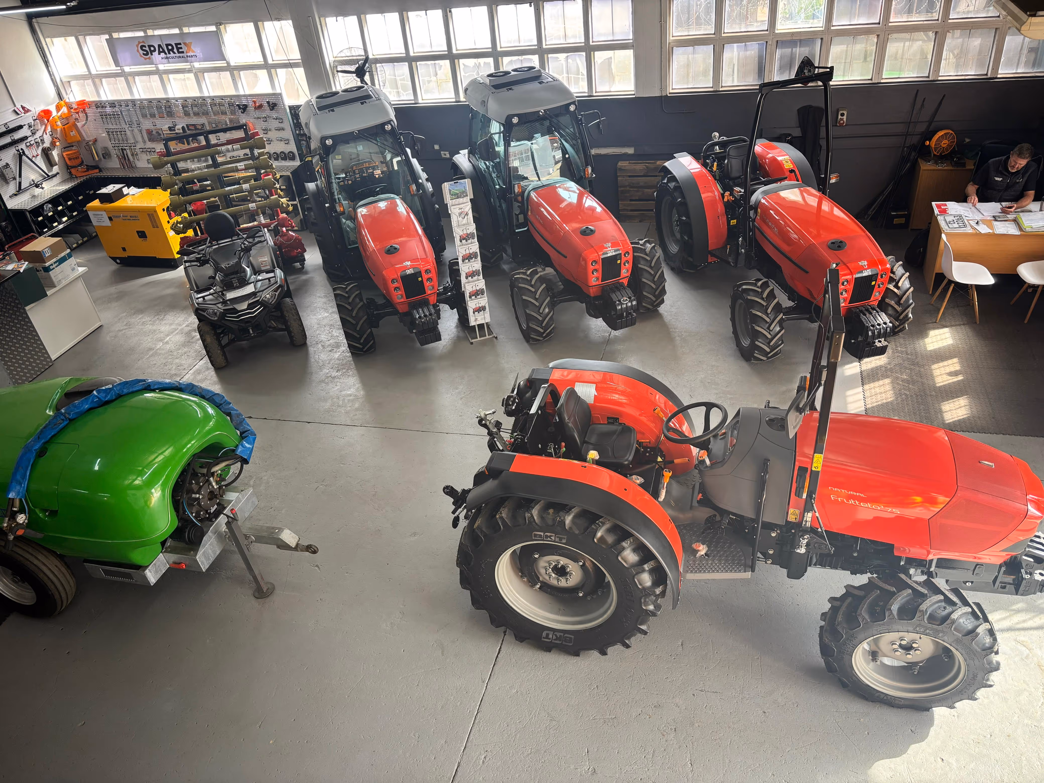 Indoor showroom with several red tractors, a green agricultural machine, and a person at a desk.