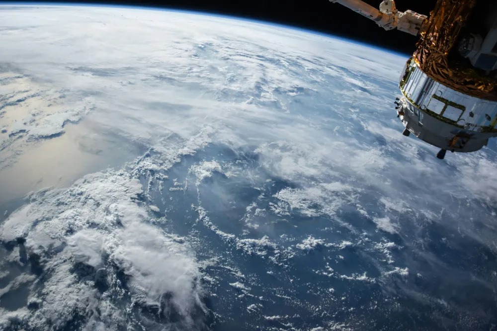 View of Earth from space showing cloud formations over the ocean with a satellite or space station module visible on the right.