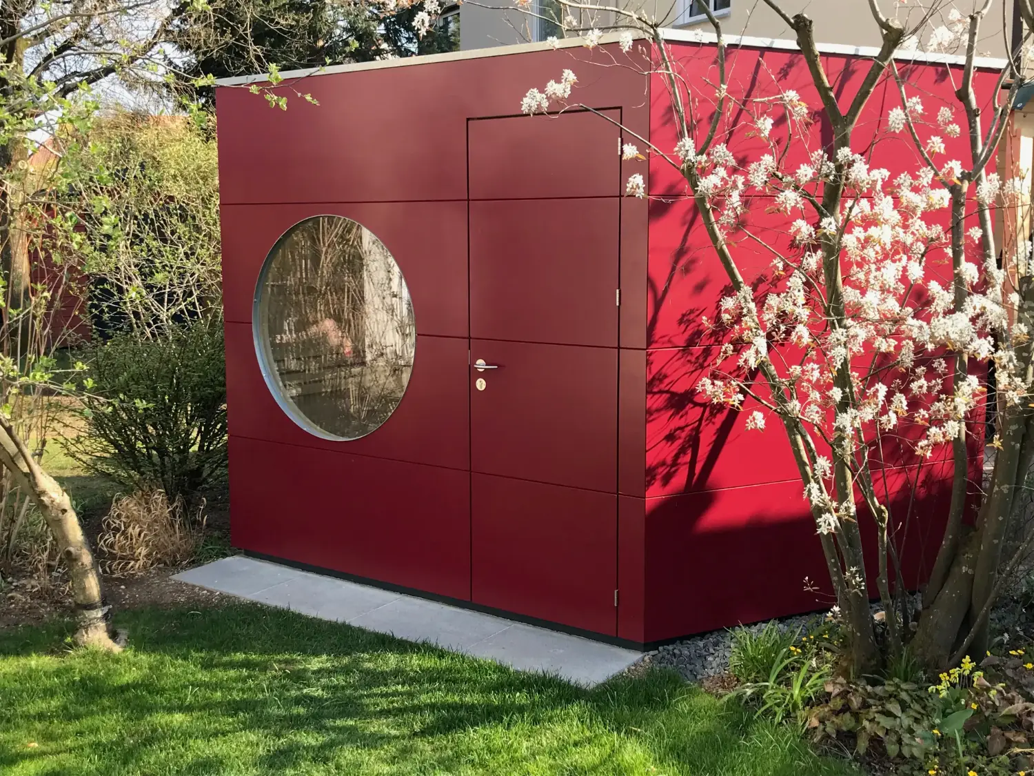 Design-Gartensauna mit roter Holzfassade und rundem Fenster, umgeben von bluehenden Straeuchern in der Fruehlingssonne