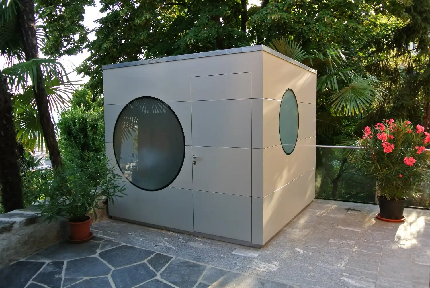 Graues Design-Geraetehaus in Kubusform mit runden Fenstern auf schoenem Steinboden vor Blumen und Palmen