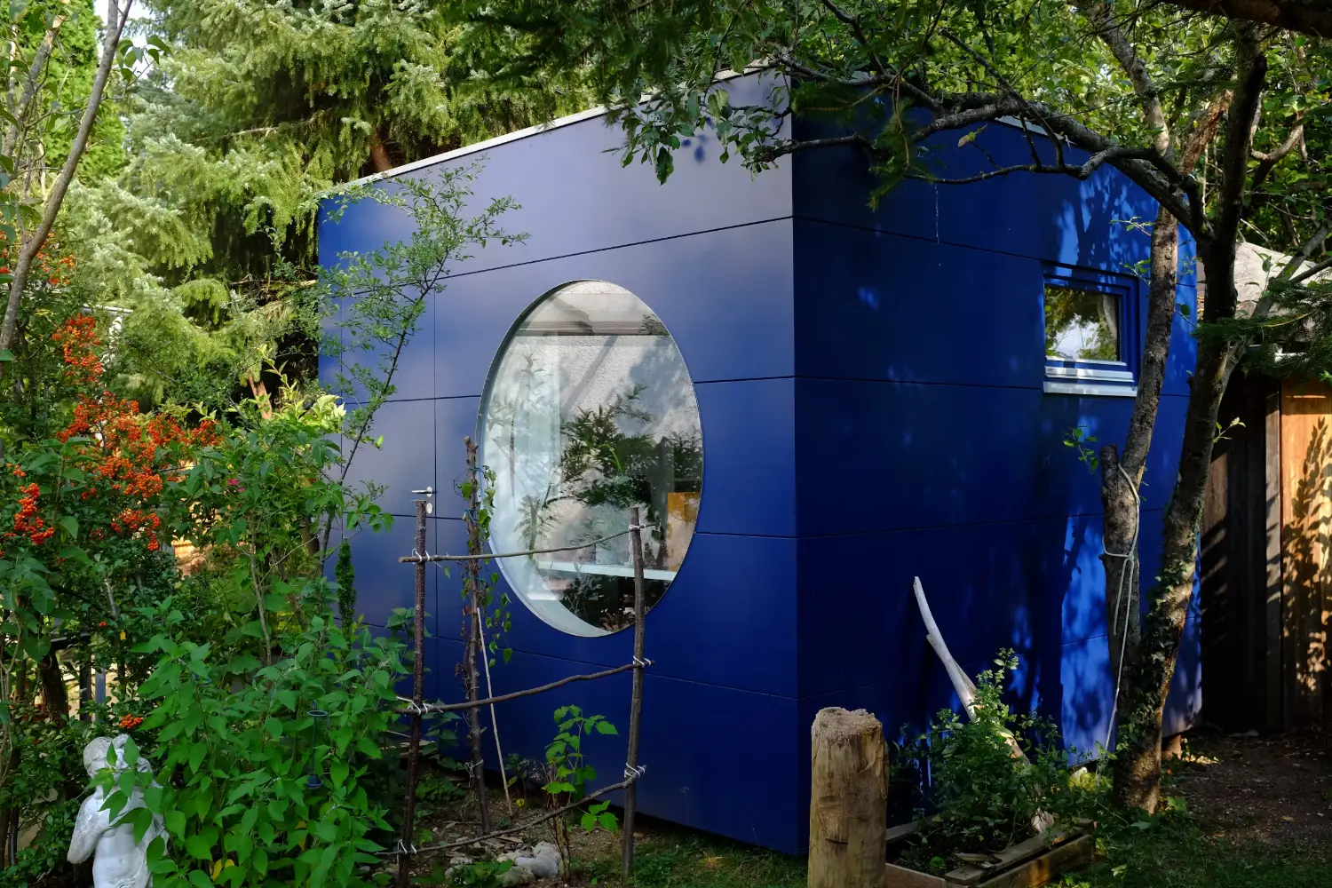 Isoliertes Gartenhaus mit blauer Holzfassade, Flachdach und rundem Fenster umgeben von gruenen Pflanzen und Baeumen