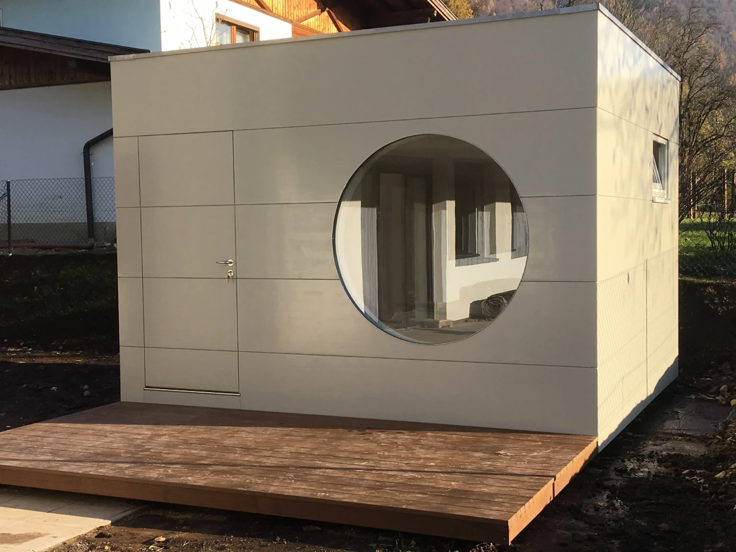 Isoliertes Design-Gartenhaus in hellem Grau mit grossem, runden Fenster, Flachdach und Holzterrasse neben Wohnhaus