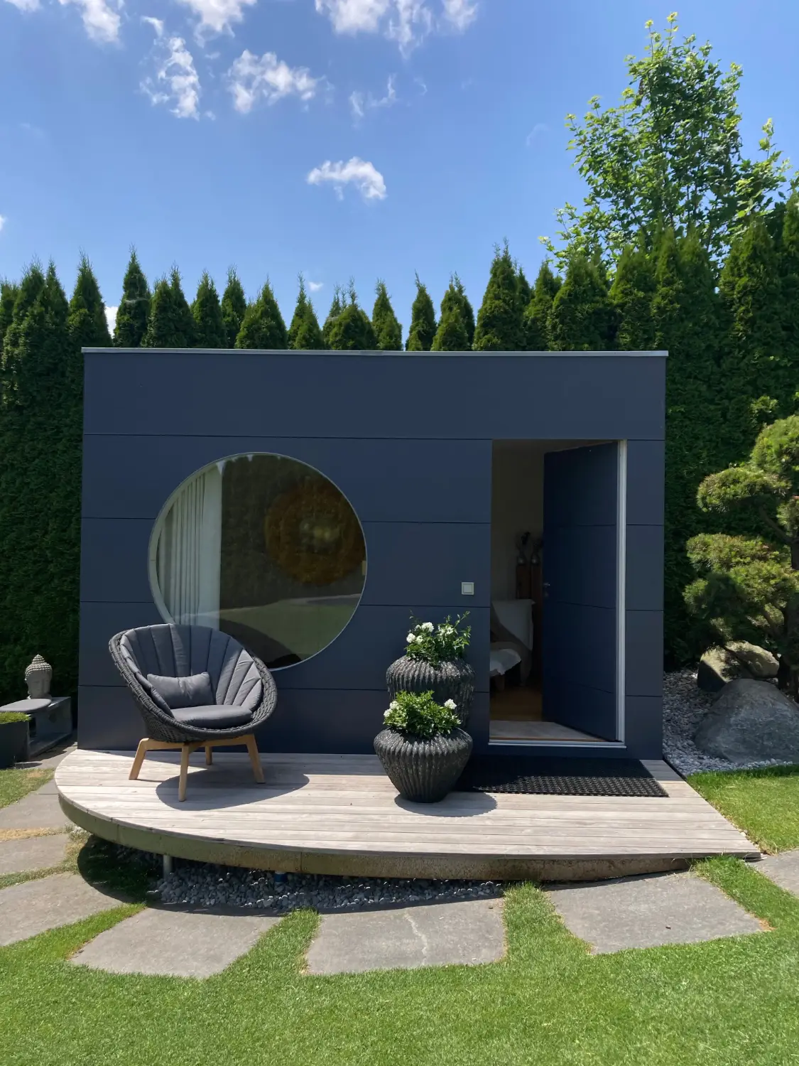 Moderner Wohnraum im isolierten Gartenhaus mit edler, dunkelgrauer Holzfassade, rundem Fenster und kleiner Terrasse vor gepflegten Hecken, umgeben von gruenem Rasen