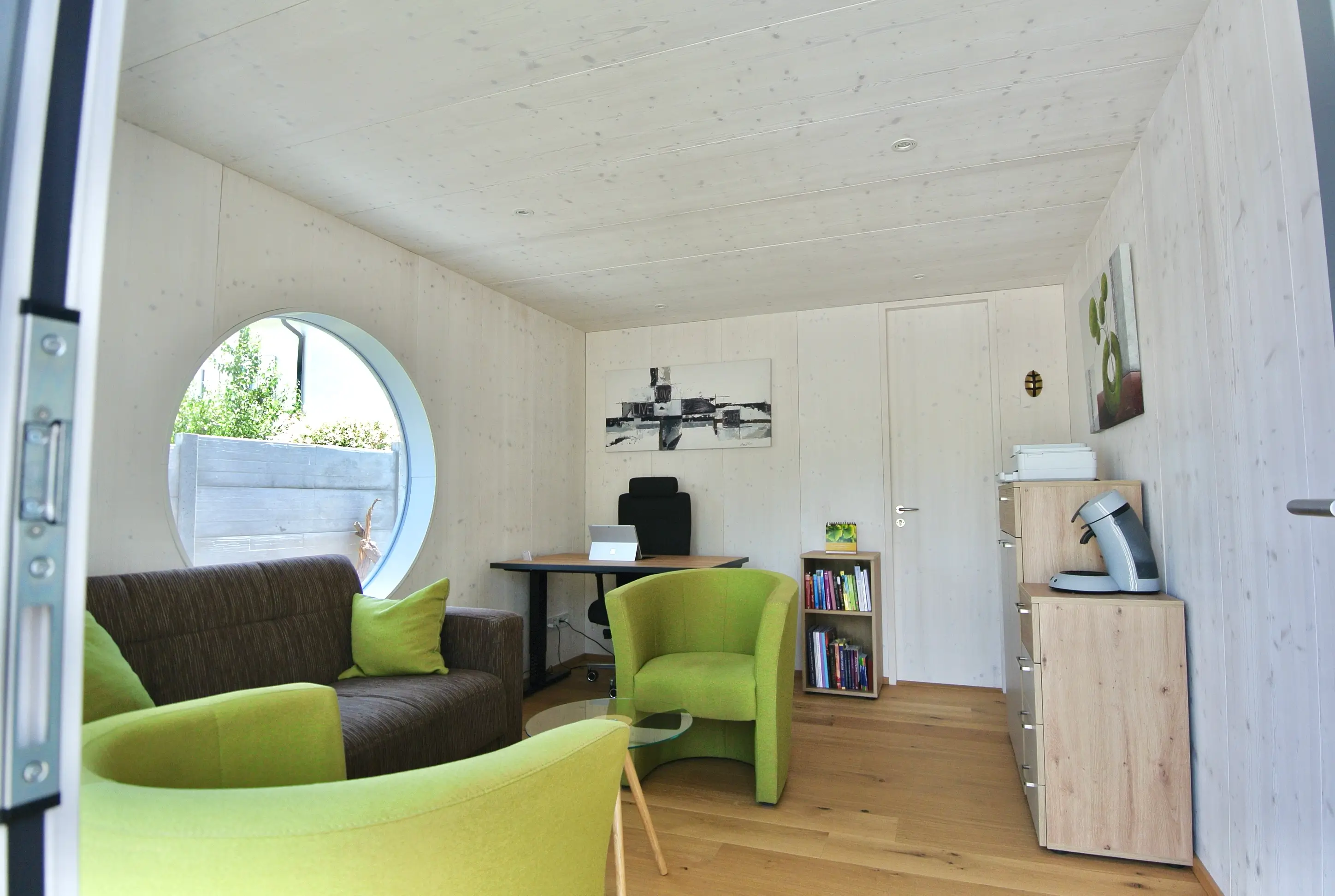Blick in den mit weiss lasiertem Holz ausgekleideten Innenraum eines isolierten Gartenhauses mit Holzboden, gruenen Sesseln und Moebeln