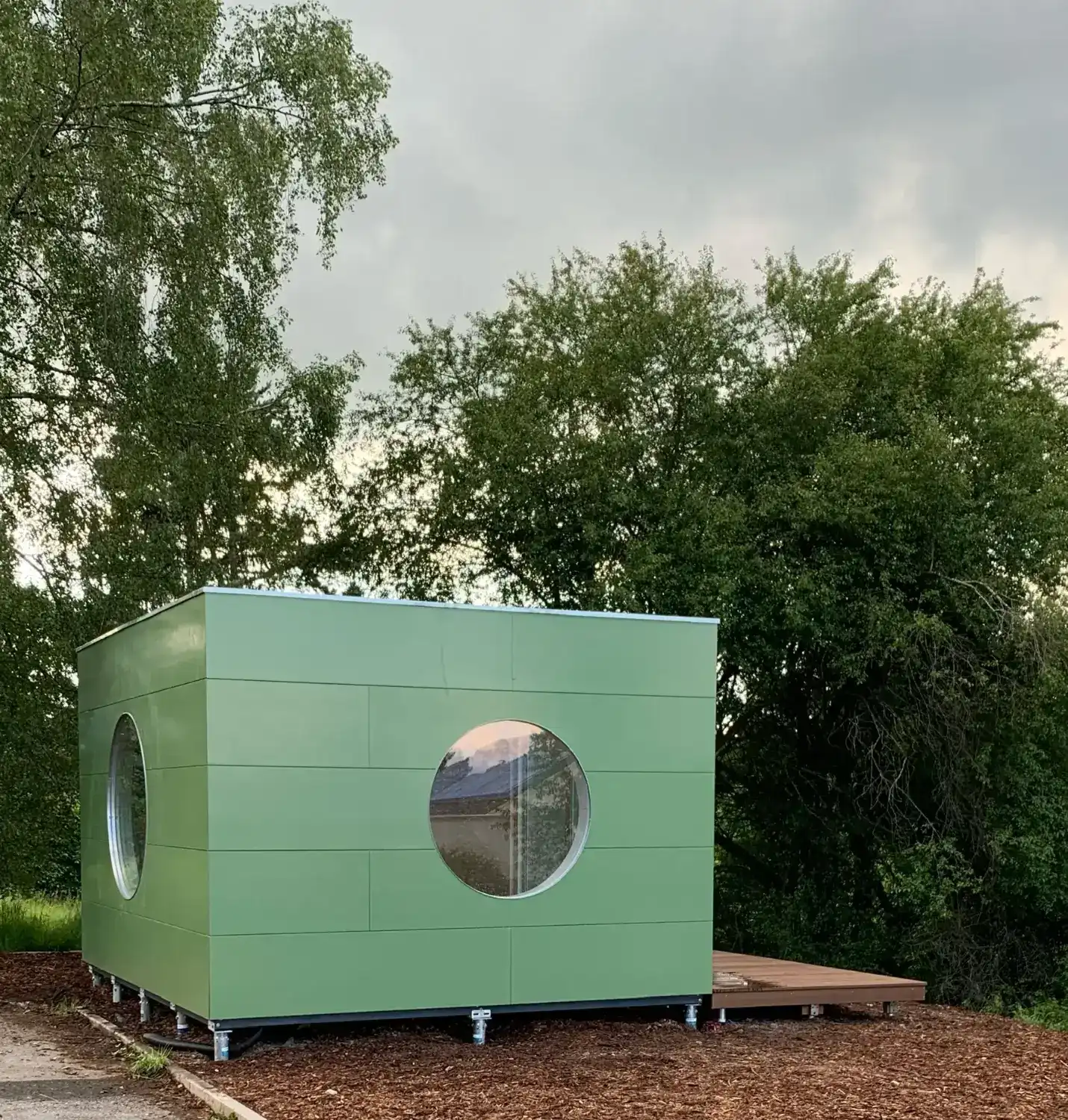 Großes, isoliertes Gartenhaus in Kubusform mit runden Fenstern und Holzterrasse umgeben von hohen, belaubten Baeumen