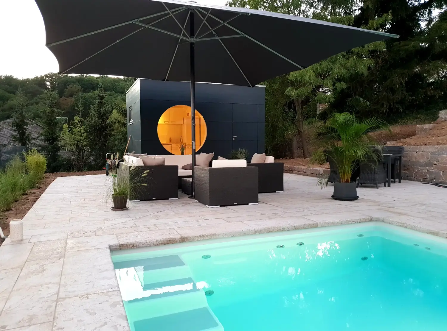 Modernes Poolhaus in Kubusform mit rundem Fenster und dunkelgrauer Holzfassade vor tuerkisem Pool, voll isoliert fuer ganzjaehrige Nutzung