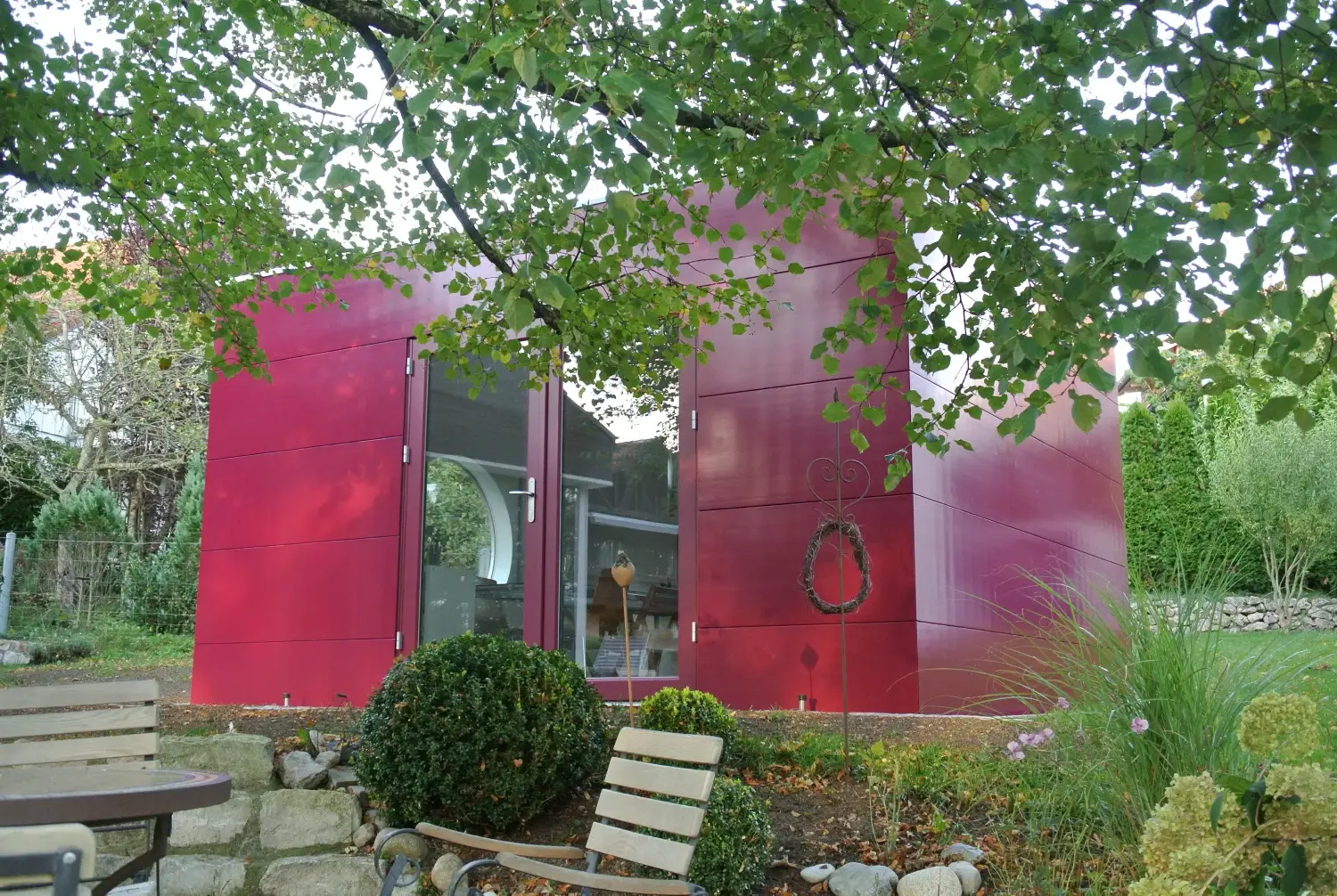 Design-Gartenhaus als Malatelier und Office mit pinker Holzfassade und verglaster Doppelfluegeltuer, stehend auf Terrasse im gruenen Garten