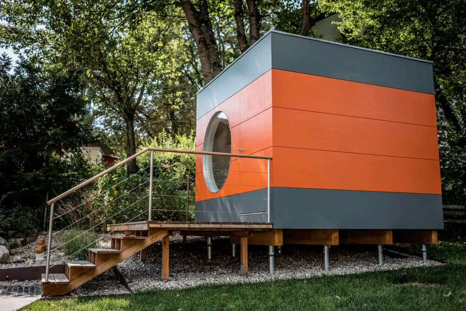 Modernes Gartenbuero mit auffaelliger Holzfassade in orange und grau, mit grosser Terrasse mit Stufen und Gelaender auf Krinnder Schraubfundamenten