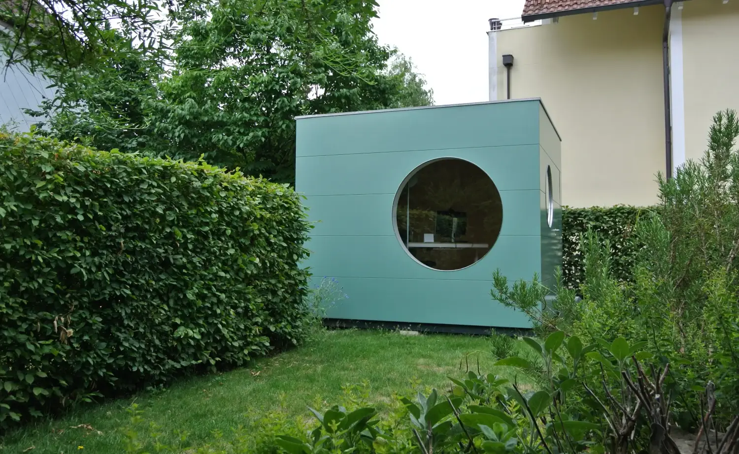 Gartenbuero mit gruenblauer Holzfassade in Kubusform mit rundem Fenster, umgeben von Hecke, gruenem Rasen und Pflanzen