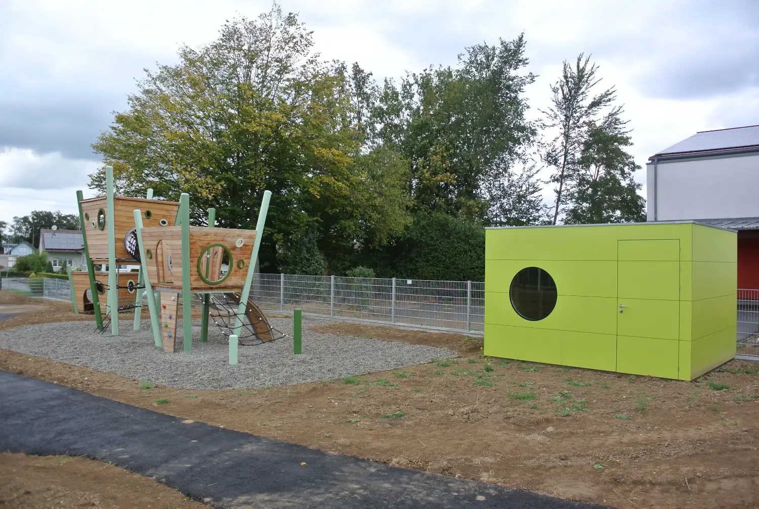Grosses, gruenes Gartenhaus als Geraetehaus eines Kindergartens, mit Flachdach und rundem Fenster, direkt neben Kletterturm
