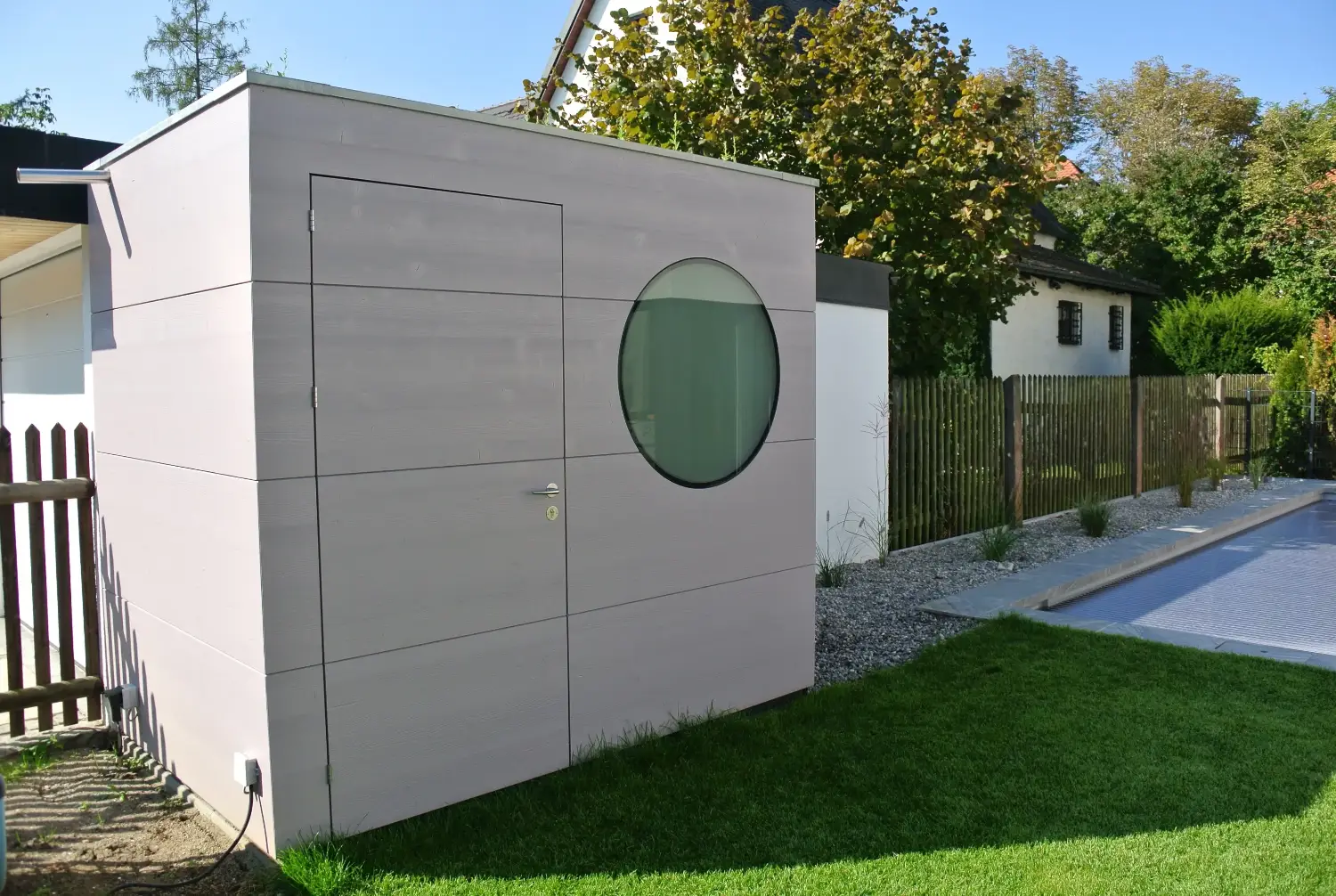 Schmales Poolhaus mit gebuersteter Holzfassade in Mauve mit rundem Fenster und Flachdach, direkt an Zaun gebaut
