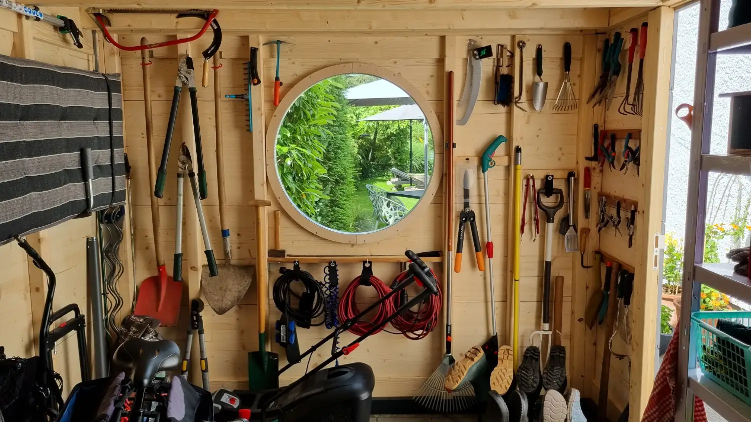 Innenraum eines Gartenhauses aus Holz mit Blick auf rundes Fenster und ordentlich organisiertem Gartenwerkzeug an den Waenden