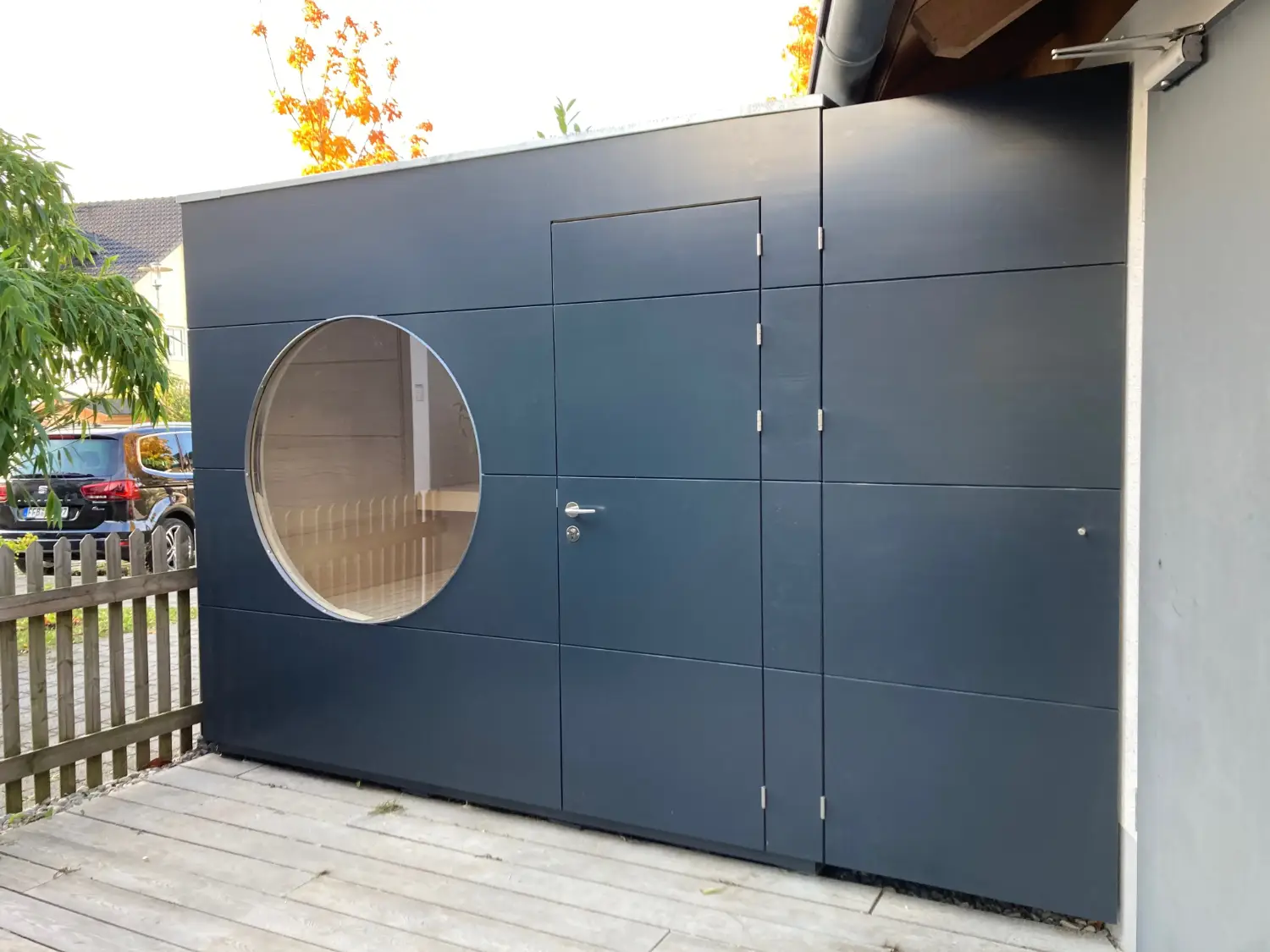 Perfekt angepasste Gartensauna zwischen Haus und Grundstuecksgrenze gebaut, mit dunkelgrauer Holzfassade und rundem Fenster