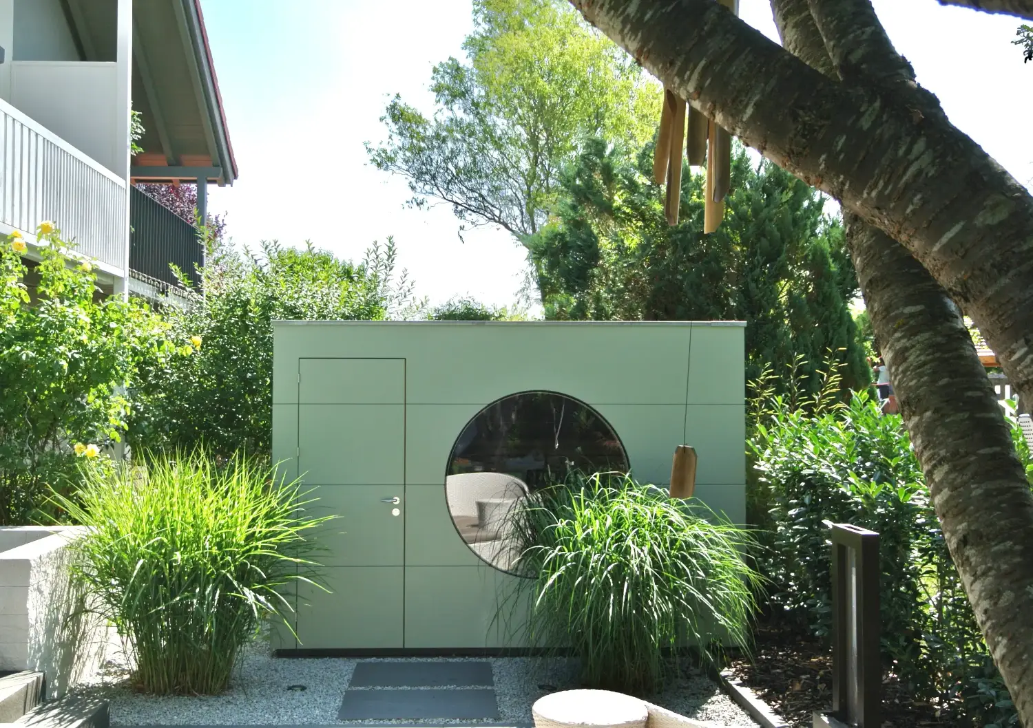 Moderne Design-Sauna im Garten mit vielen Pflanzen und Baeumen, die gruene Holzfassade verschmilzt harmonisch mit der Umgebung