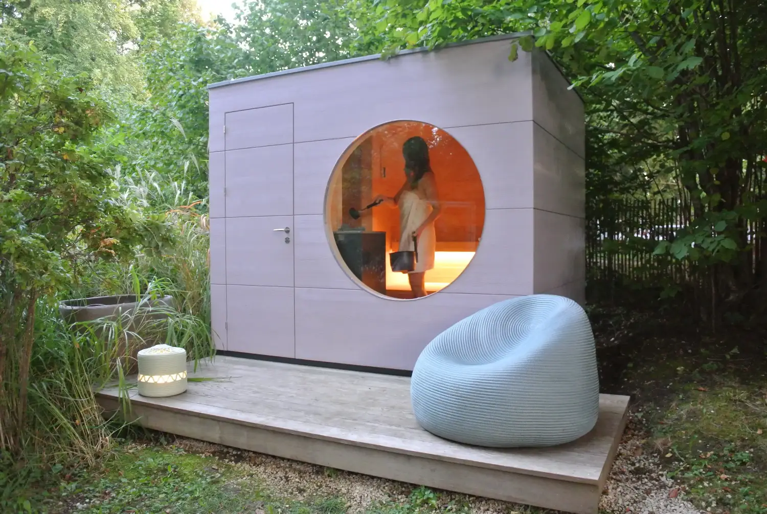 Gartensauna umgeben von Baeumenmit Holzfassade in Mauve und Holzterrasse, durch das runde Fenster sieht man Frau im Inneren Aufguss machen