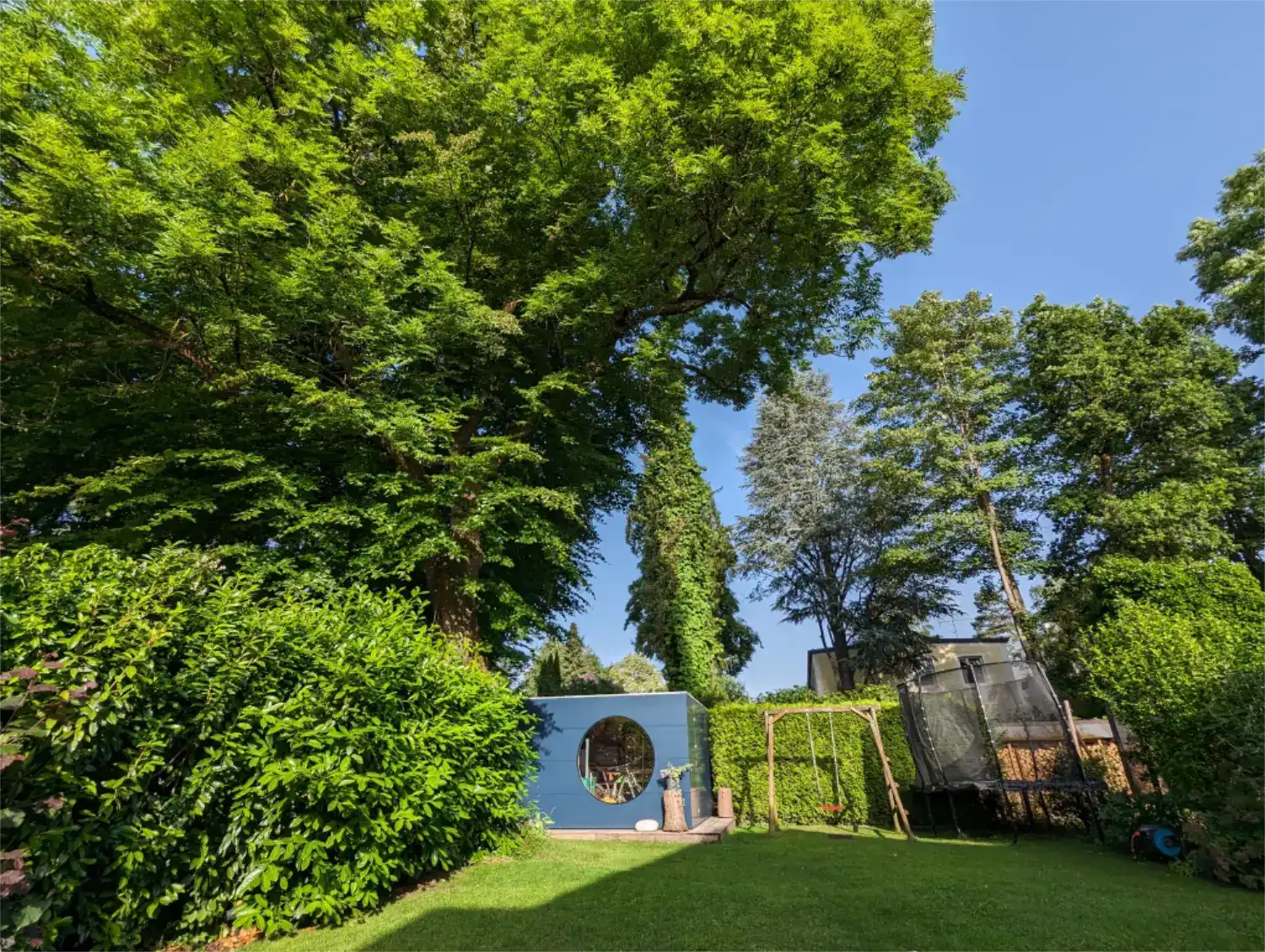 Großer Garten mit sattgruenen Baeumen, Straeuchern, Wiesen, mittendrin steht ein kubusfoermiges Design-Gartenhaus in dunkelgrau mit rundem Fenster
