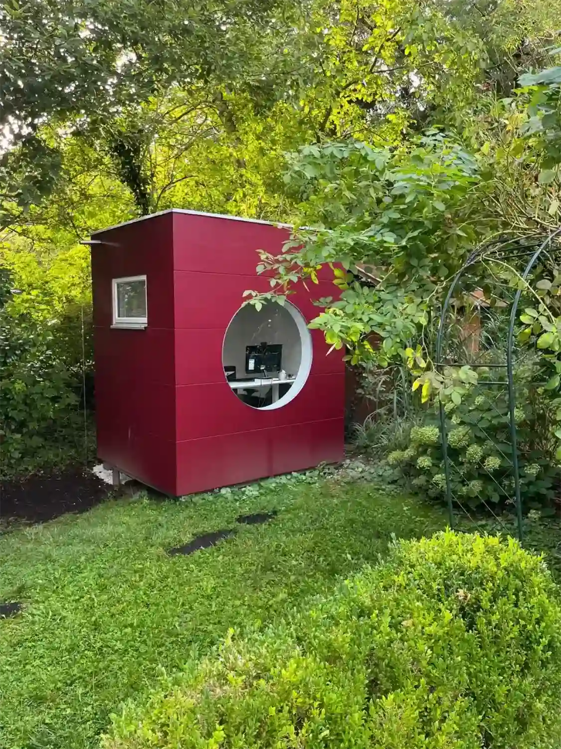 Blick auf ein kleines Homeoffice fuer eine Person mit rundem Fenster, Flachdach und roter Holzfassade im gruenen Garten