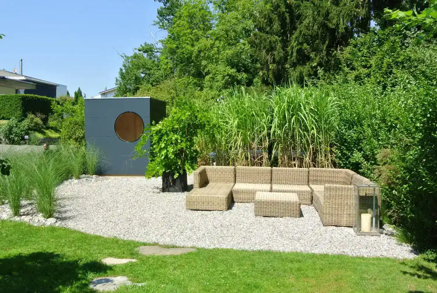 Großer Garten mit Sitzlounge, umgeben von sattgrünen Pflanzen, daneben ein anthrazitfarbenes Design-Gartenhaus in Kubusform