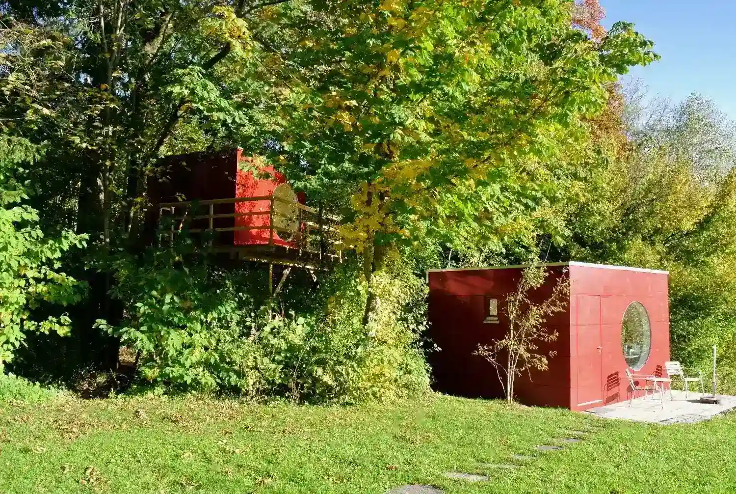 Blick auf 2 isolierte Gartenhaeuser im gruenen Garten mit roter Holzfassade, ein Kubus steht erhoeht in den Baeumen mit Balkon