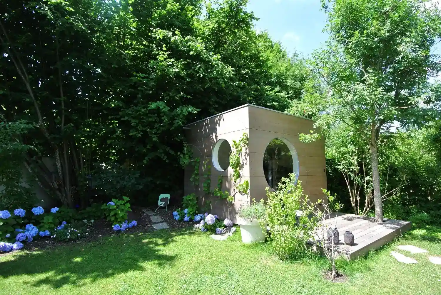 Neuroaesthetisch in gruenen Garten mit blauen Blueten integriertes Kubus Gartenhaus mit runden Fenstern, Holzfassade