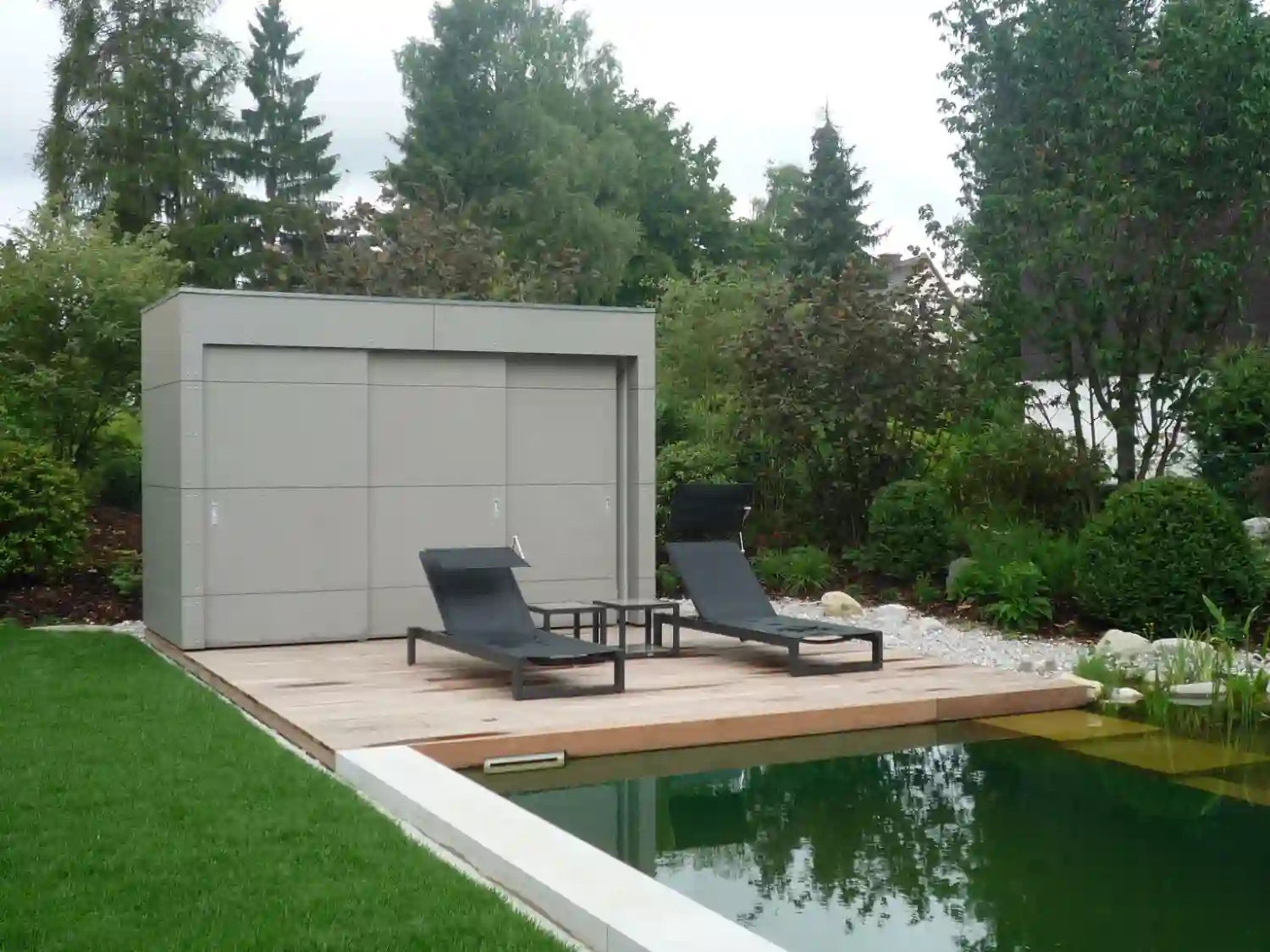 Design Poolhaus mit grauer Eternitfassade, Flachdach, Schiebetueren, davor Holzterrasse und Naturpool