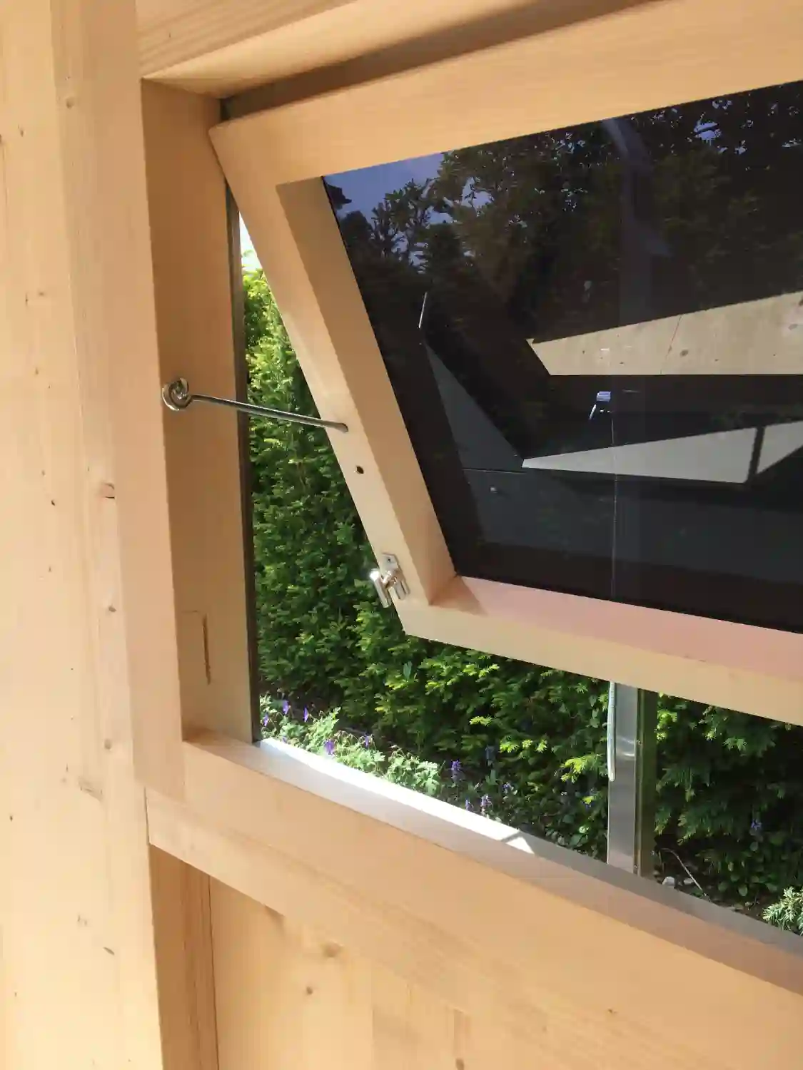 Detailblick auf Lüftungsfenster aus Holz mit schwarzer Scheibe eines Gartenhauses, Fenster gekippt, Blick auf Hecke
