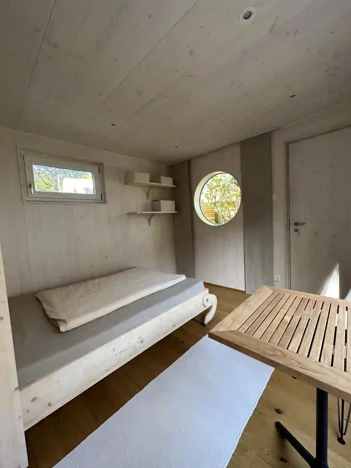 Im Innenraum eines Tiny Houses im Gartenhaus mit Eicheparkett, weiß lasierten Holzwaenden, Tisch, Klappbett, Rundfenster