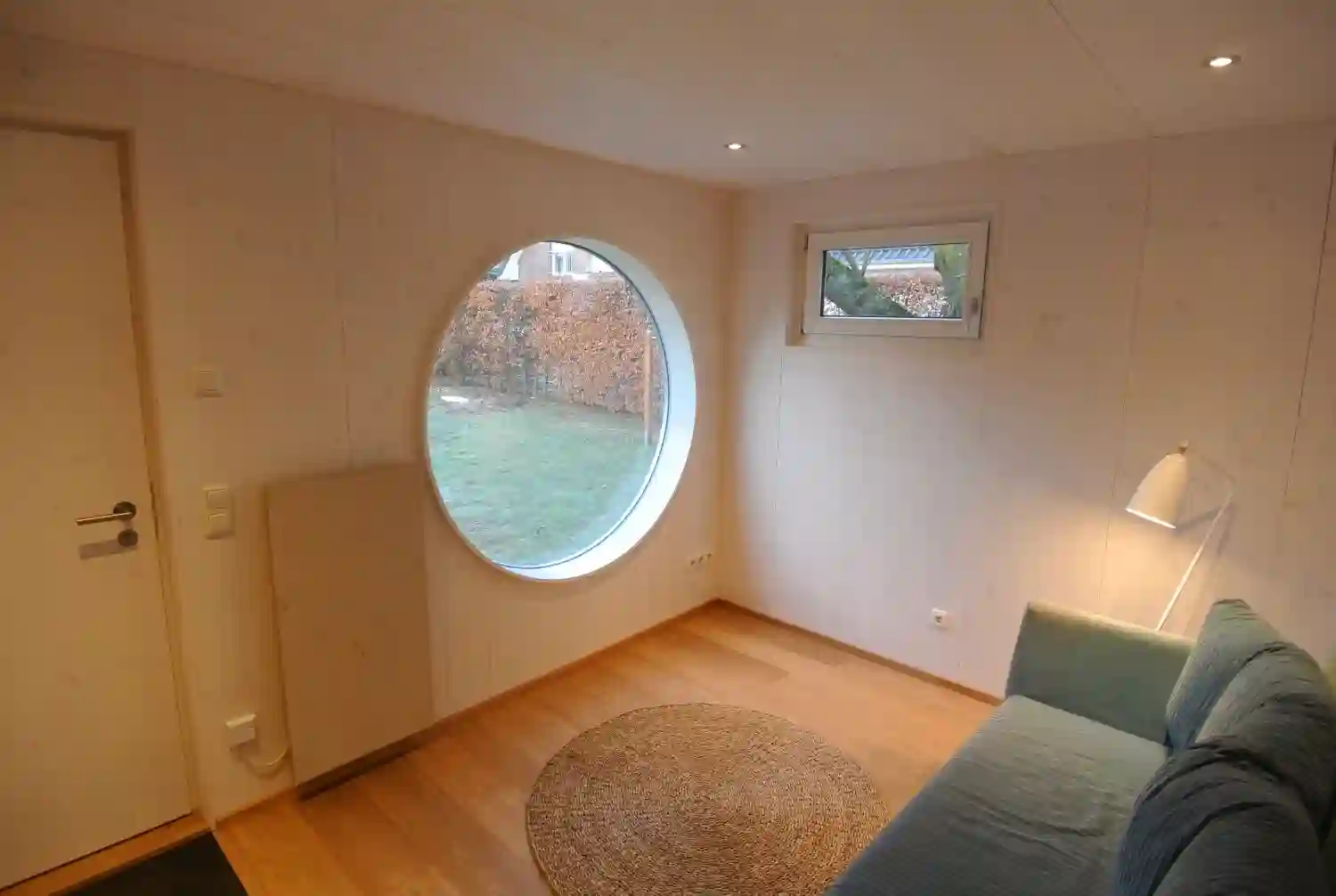 Im Wohnraum eines isolierten Garten Kubus Gartenhauses mit Rundfenster, Lüftungsfenster, Tuer, Heizstein, gruenem Sofa