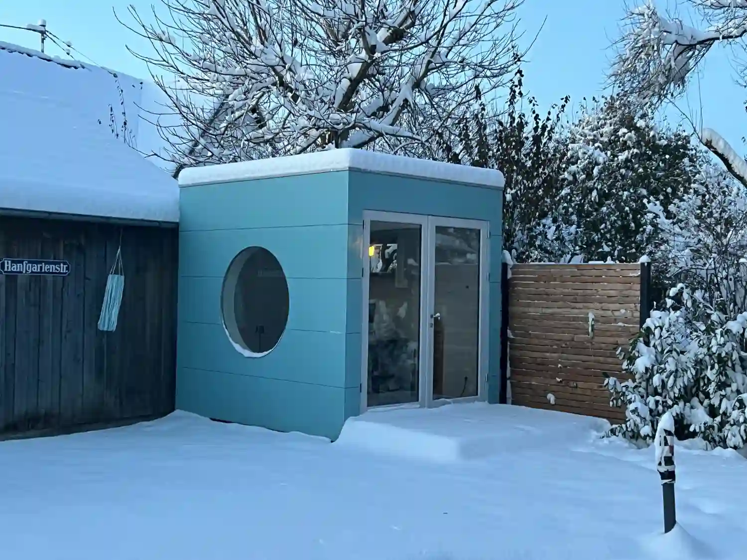 Modernes Gartenbüro mit Rundfenster, blau gestrichener Holzfassade im verschneiten Garten mit Schnee auf Flachdach