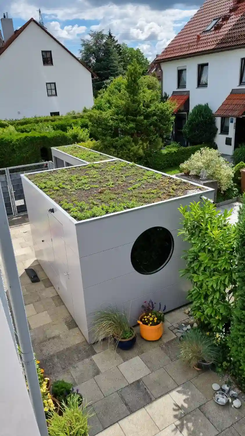 Modernes Garten Kubus Gerätehaus mitten in Wohnsiedlung mit grau gestrichener Holzfassade, Rundfenster, begrüntem Flachdach
