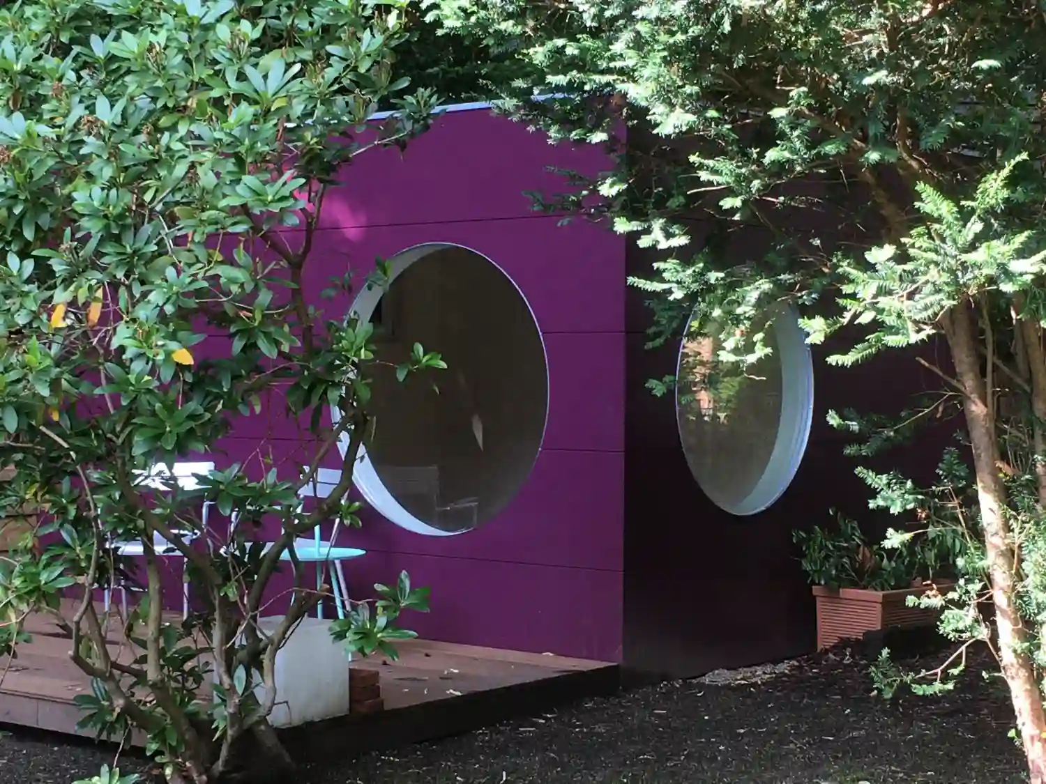 Design Gartenhaus mit lila gestrichener Holzfassade, Flachdach, 2 Rundfenstern, Holzterrasse umgeben von grünen Bäumen