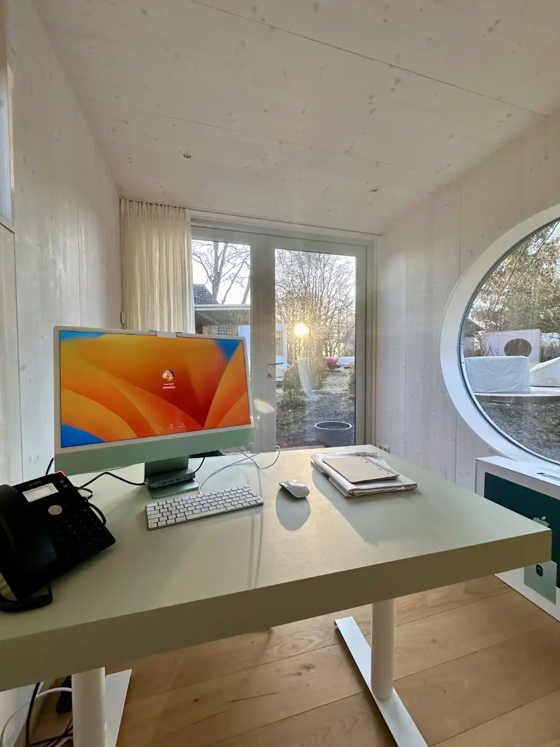 Im Innenraum eines Gartenbüros mit Blick aus verglaster Doppelflügeltür und rundem Fenster in winterlichen, sonnigen Garten