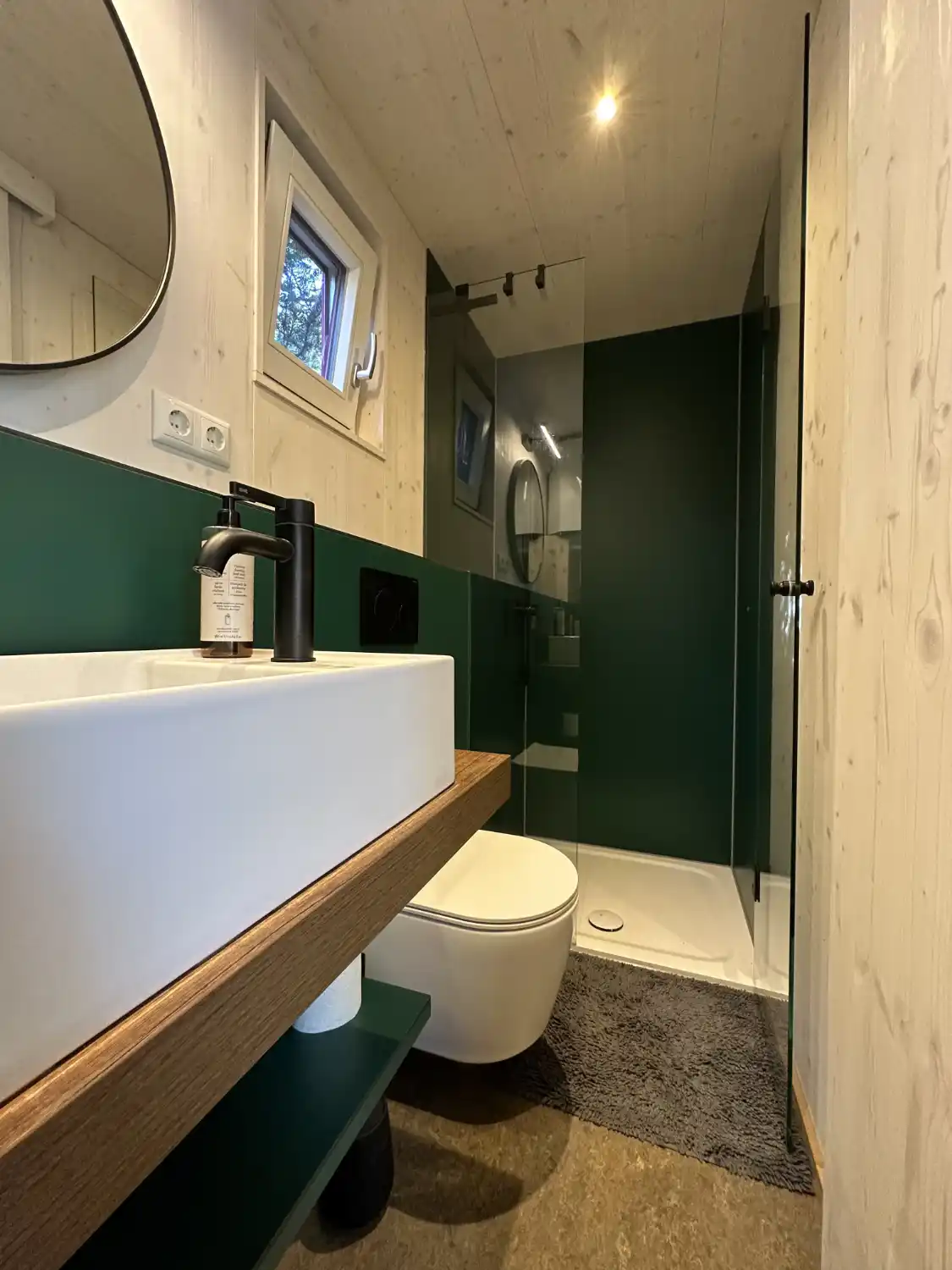 Blick ins kleine Bad mit WC und Dusche mit weiß lasierten Holzwänden und grünen Details eines Tiny House Gartenhauses