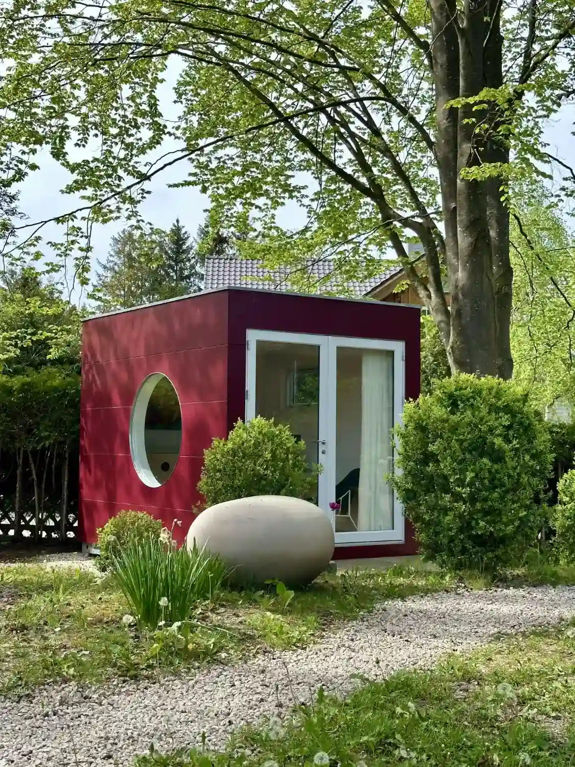 Design Gartenbüro mit Flachdach, Doppelflügeltür, Rundfenster und rot gestrichener Fassade umgeben von Bäumen und Büschen