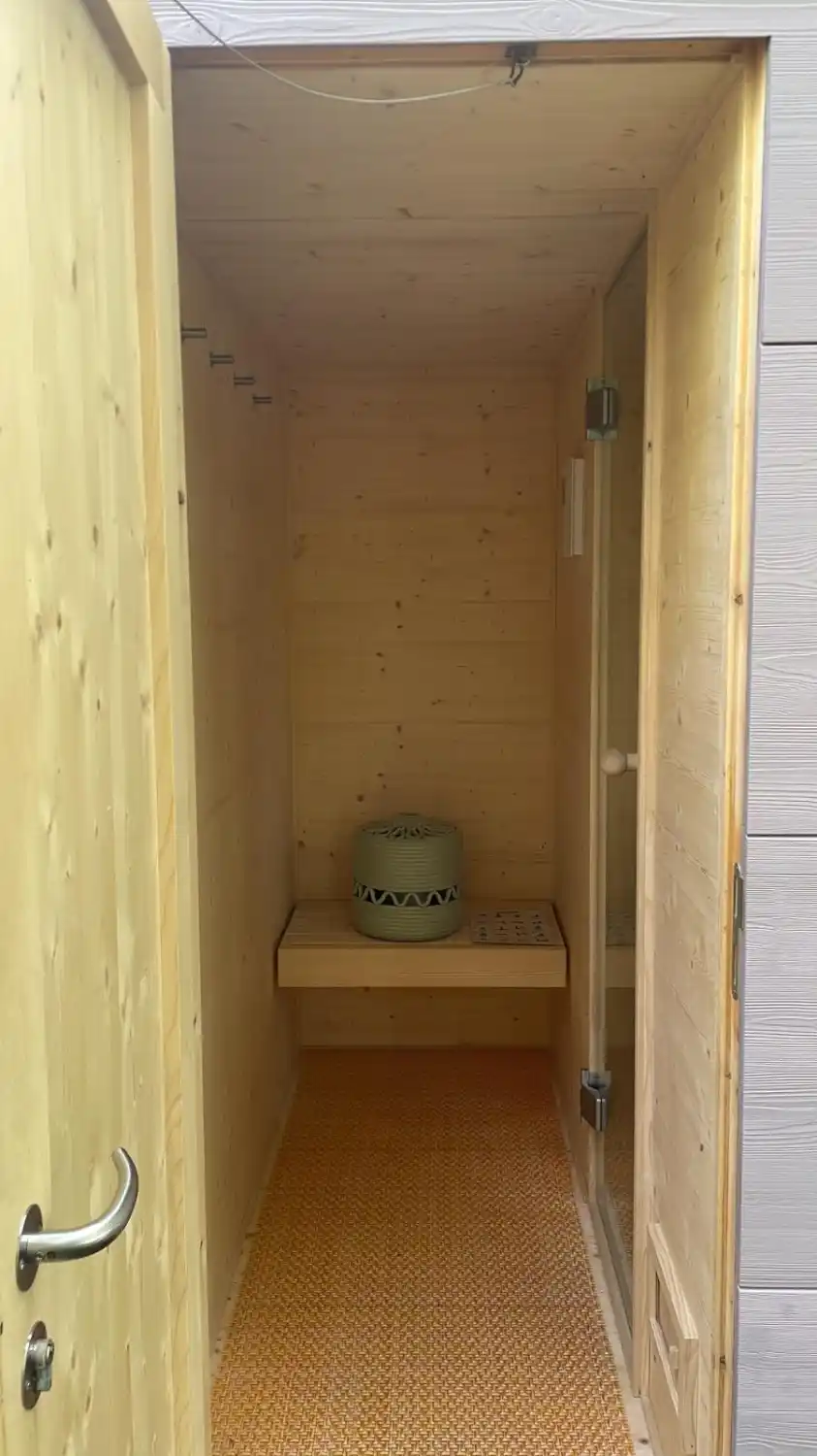 Blick durch offene Tuer einer Gartensauna in den mit Holz ausgekleideten Vorraum, gegenueber Sitzbank, links Kleiderhaken, rechts Glastuer zu Saunaraum