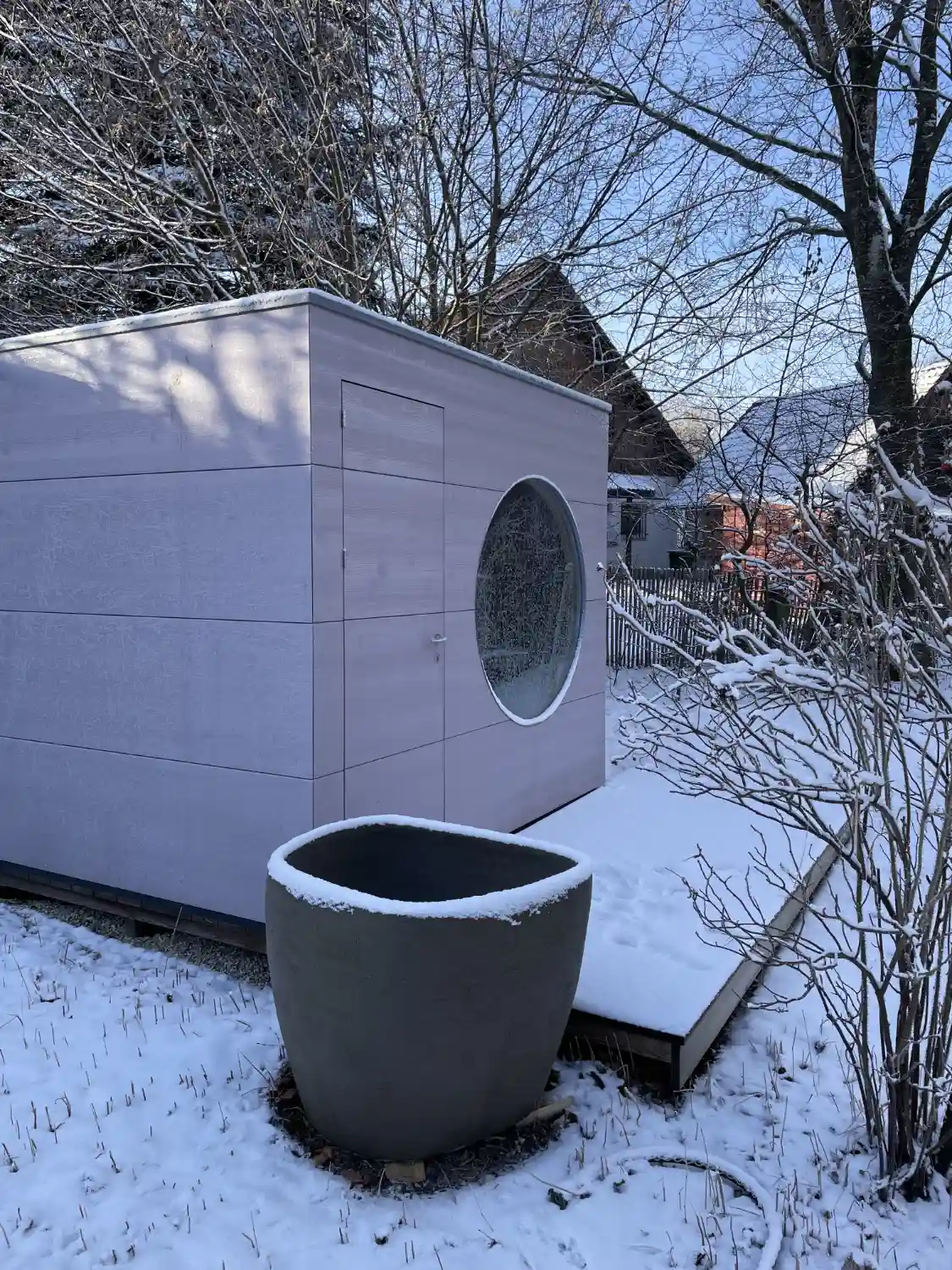 Blick auf moderne Gartensauna in Kubusform mit Rundfenster und gebuersteter Holzfassade aus Fichte im verschneiten Garten