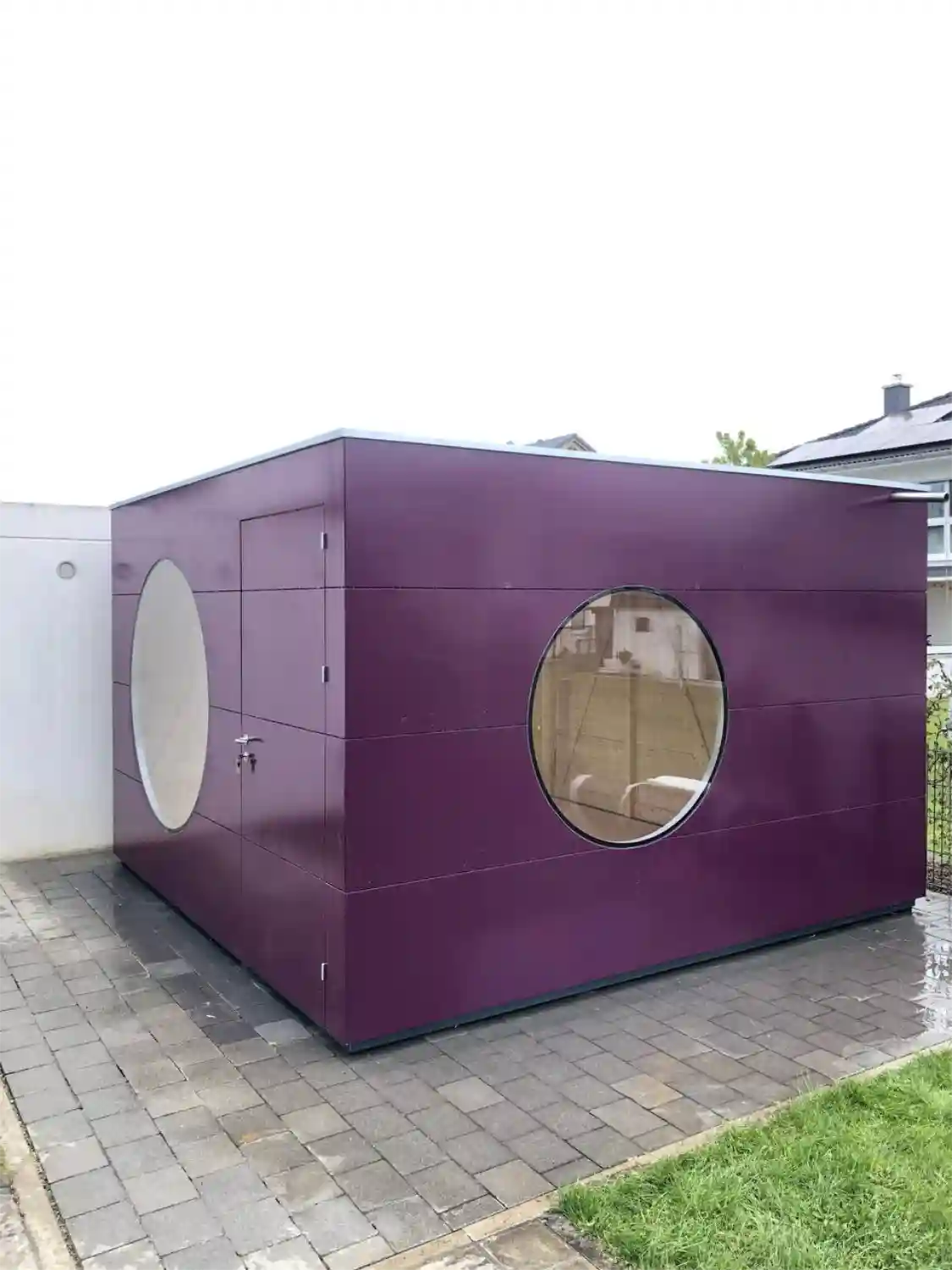 Gartenhaus-Kubus mit Rundfenstern und lila gestrichener Holzfassade, vom nassen Boden geschuetzt dank Alurahmen