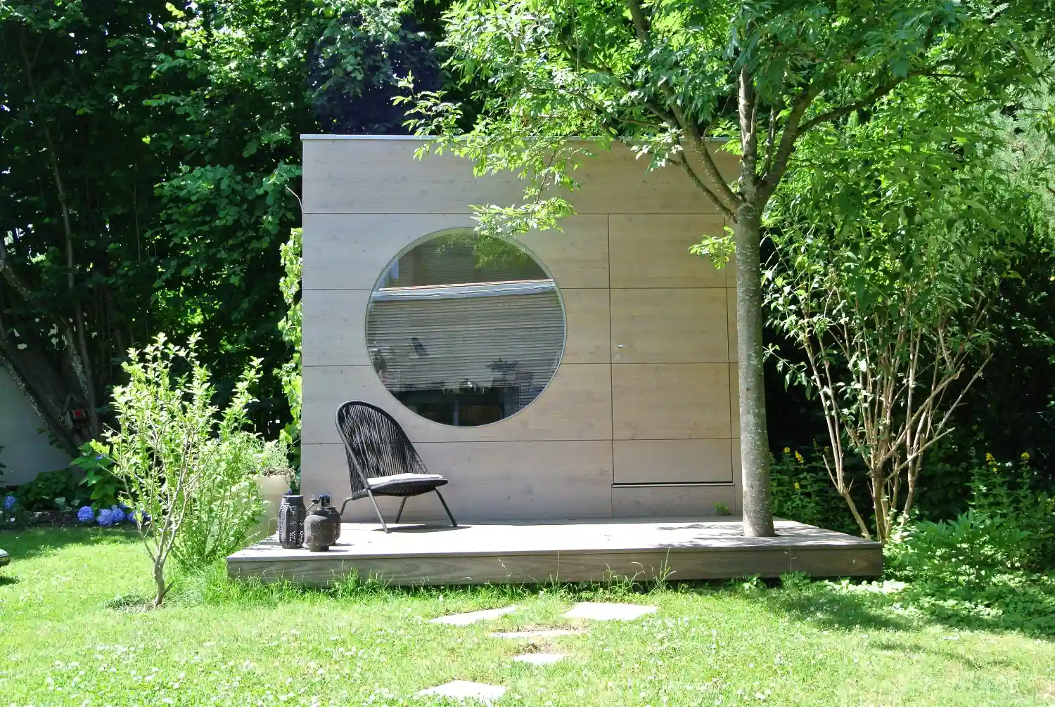 Kubusfoermiges Design Gartenbuero mit Rundfenster, Terrasse und Holzfassade aus Laerche natur lasiert mit Vergrauungslasur