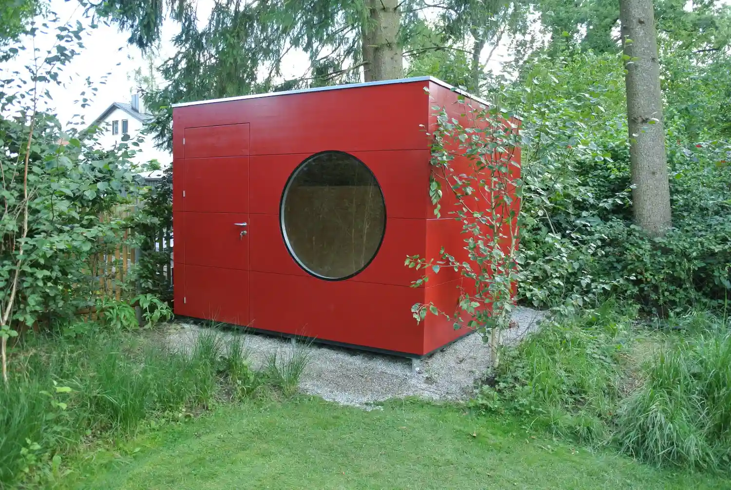 Rotes Gerätehaus mit Rundfenster und Flachdach zwischen sattgrünen Pflanzen, auf gekiester, wasserdurchlässiger Fläche
