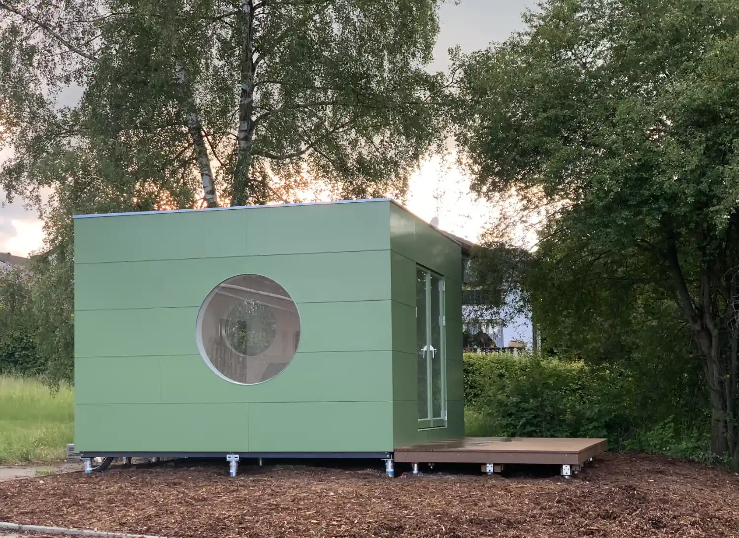Wohn Kubus mit gruener Holzfassade und Terrasse, montiert auf Schraubfundamenten, im Hintergrund Hecke, Baeume und Wohnhaus