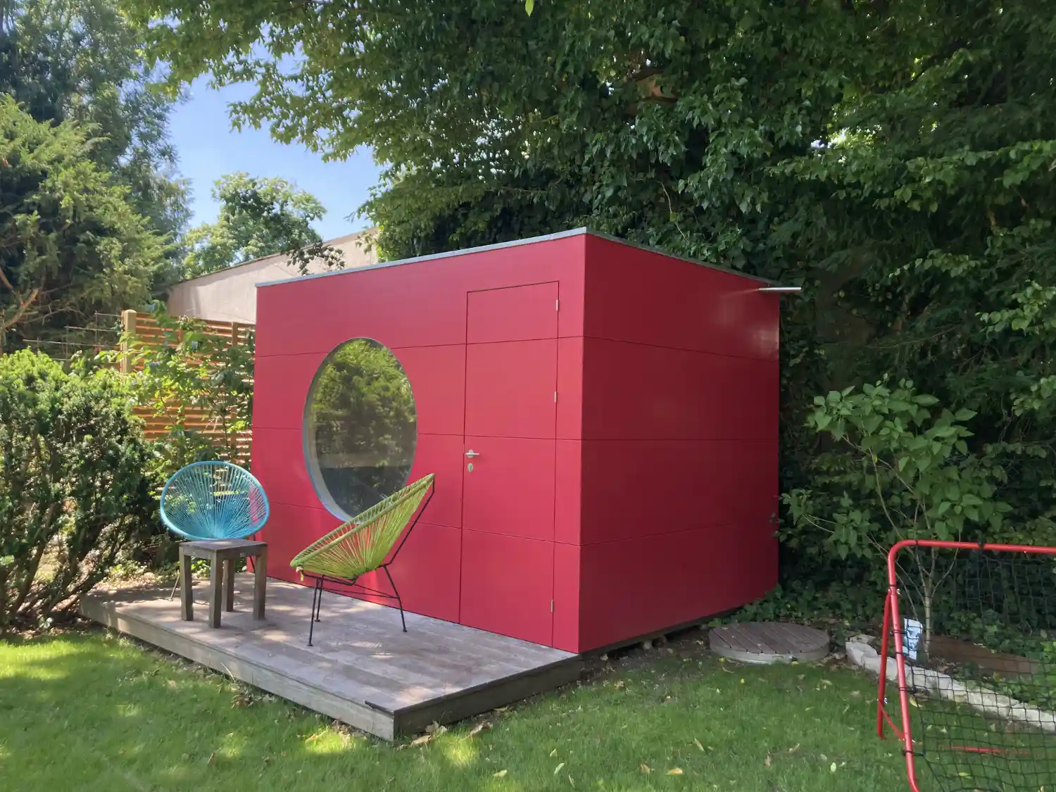 Rote Gartensauna mit Flachdach, Rundfenster und Holzterrasse auf Schraubfundamenten, dahinter gruene Baeume