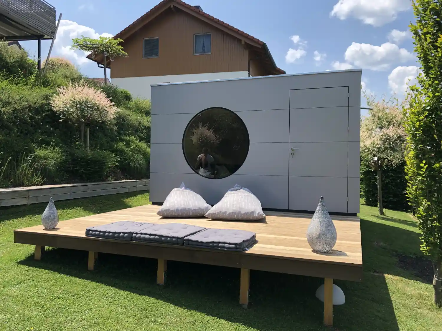 Graues Design-Gartenhaus mit Rundfenster und grosser Holzterrasse, zum Ausgleich der Hanglage auf Schraubfundamenten gebaut