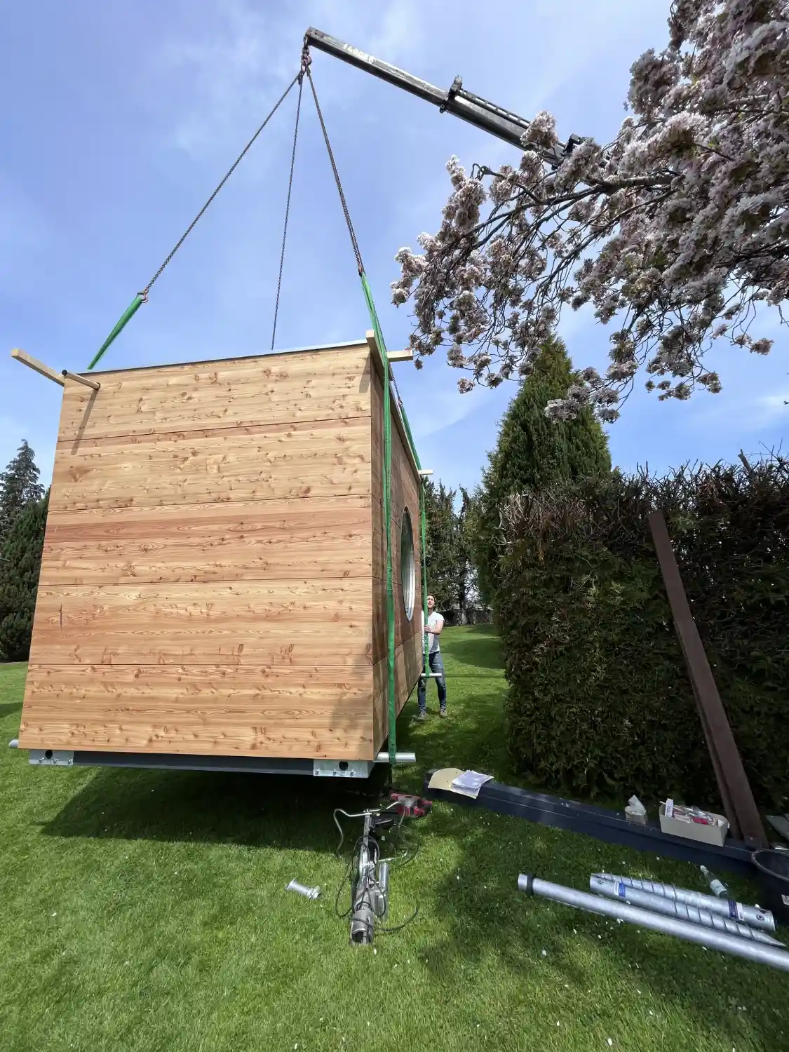 Gartenhaus mit Laerche-Fassade haengt an einem Kran und wird in einen Garten gehoben zur Montage auf Schraubfundamenten