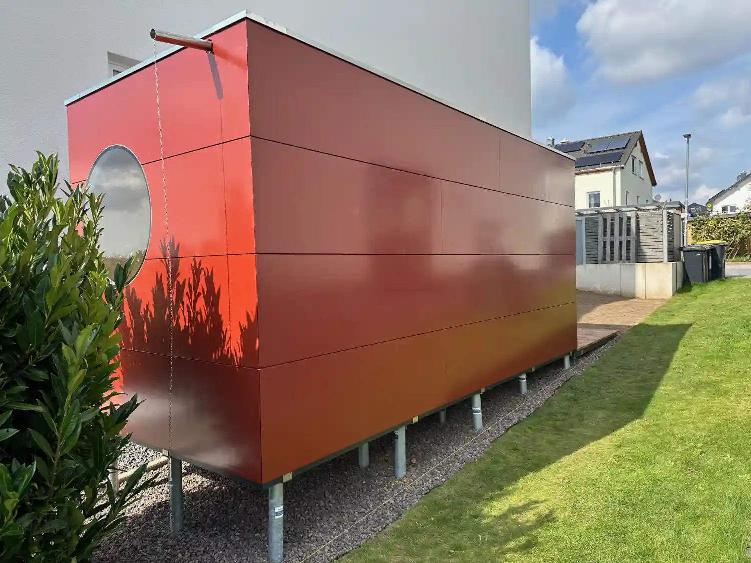 Längliches rotes Gartenhaus auf Schraubfundamenten zwischen zwei Häusern in leichter Hanglage