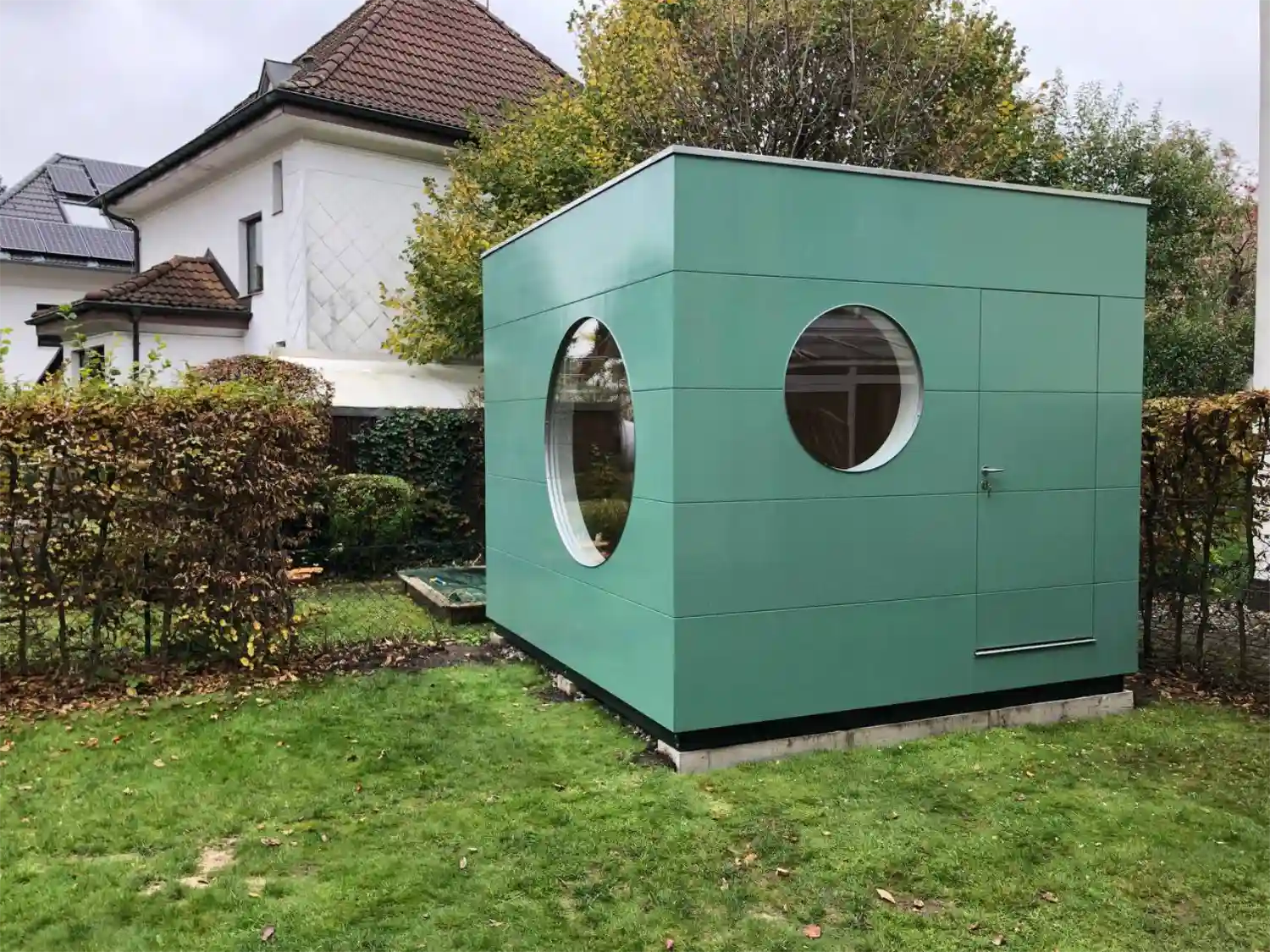 Grünes Gartenbüro mit zwei Rundfenstern und Flachdach auf sichtbaren Streifenfundamenten, im Hintergrund Wohnhäuser und Bäume