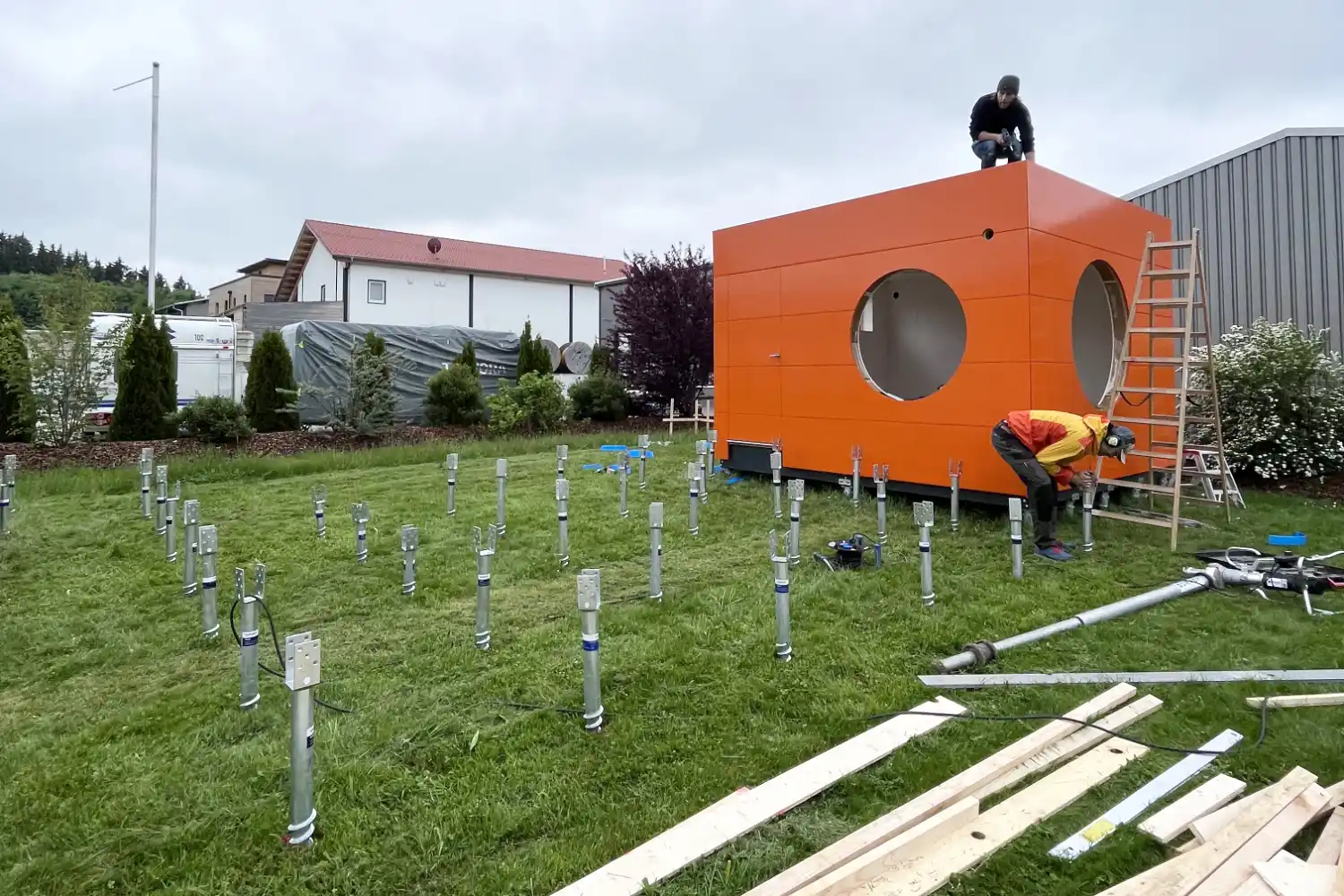 Orangener Office Kubus bei der Montage auf Krinner-Schraubfundamenten, davor weitere Schraubfundamente für die Terrasse