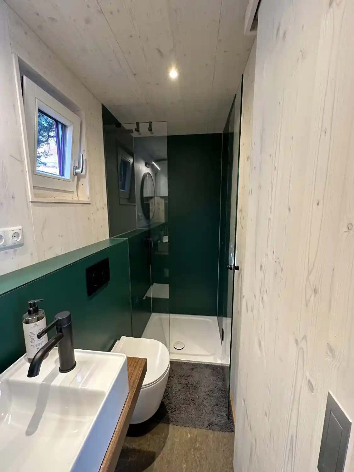 Separater Badbereich des Tiny Houses mit Dusche, Glastür, WC und Waschbecken, weiß lasierten Fichtenwänden und grünem Linoleum