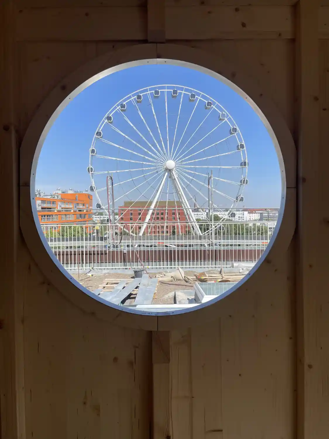 Ausblick auf das Münchner Riesenrad durch das Rundfenster eines Gartenhauses, dass auf einer Dachterrasse montiert ist.